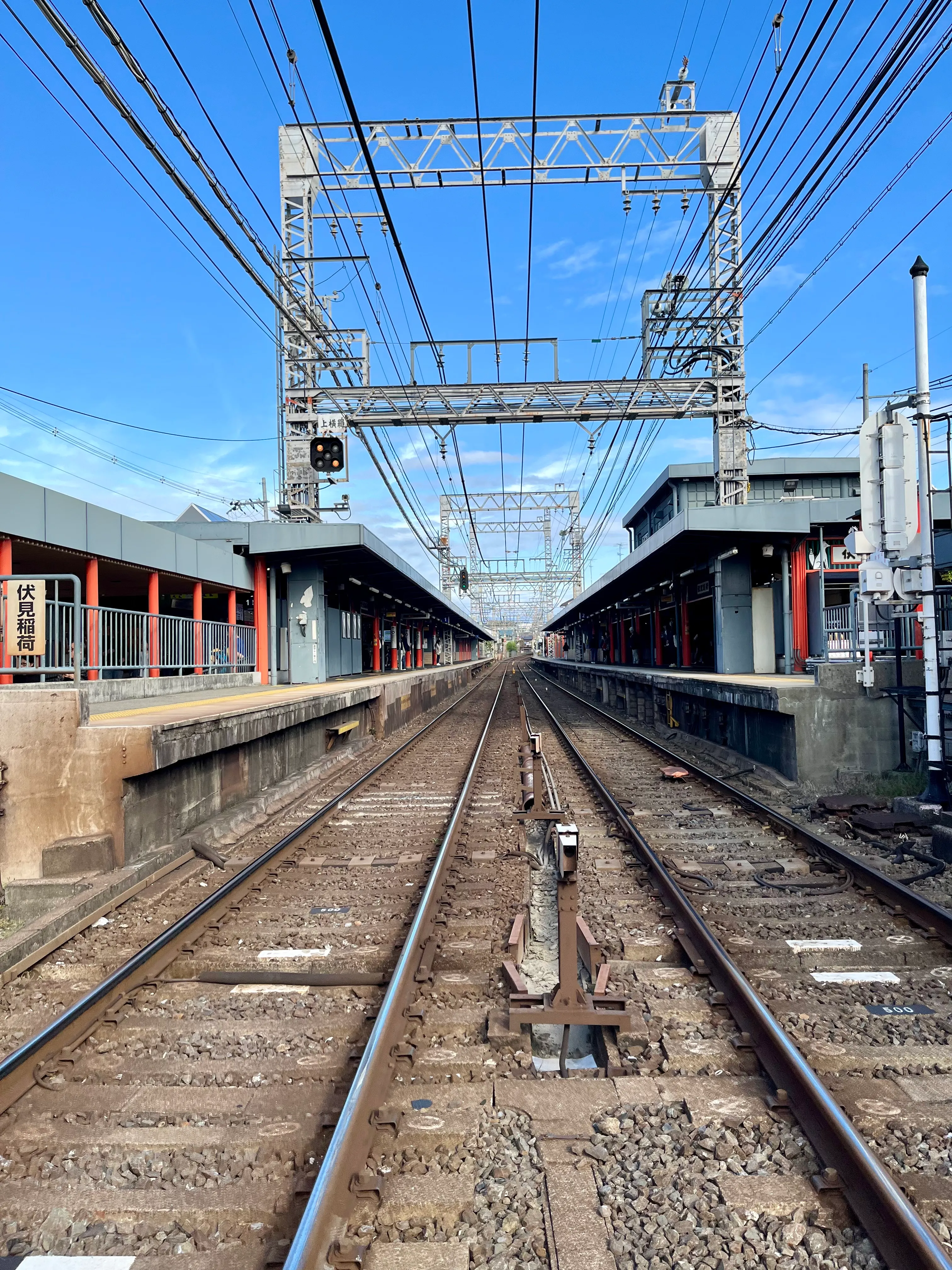 A pair of train tracks and overhead wires run straight past a platform and into the distance.