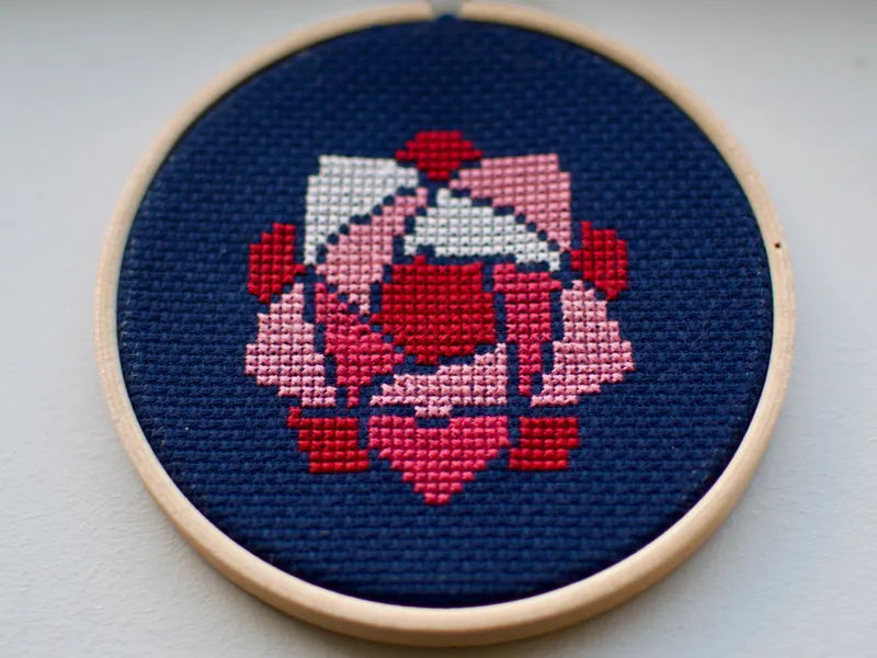 A small cross-stitch of a pink, roselike flower against a navy background.