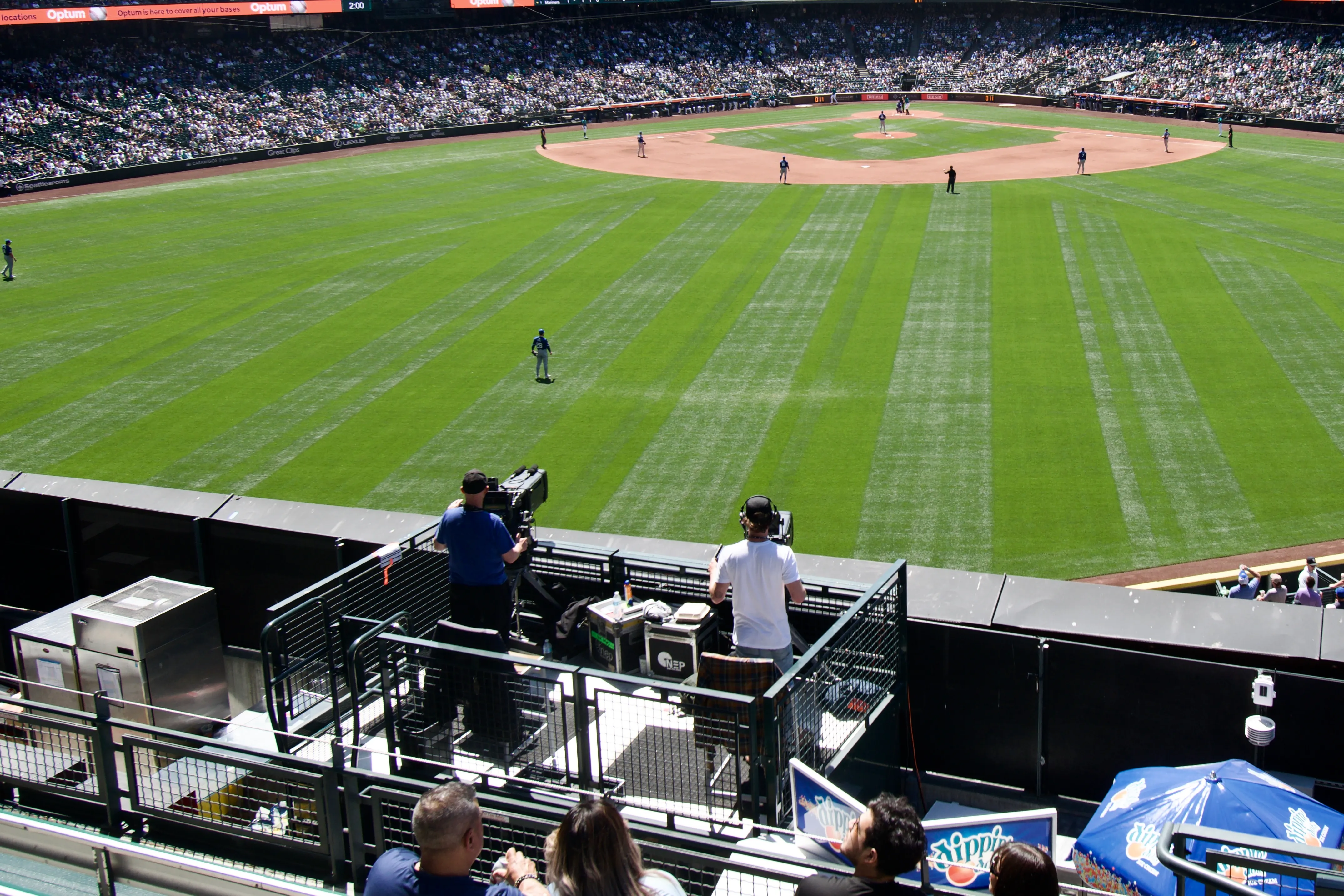 Two camera operators stand on a fenced-in platform above the center-field wall and point their cameras at the pitcher and batter