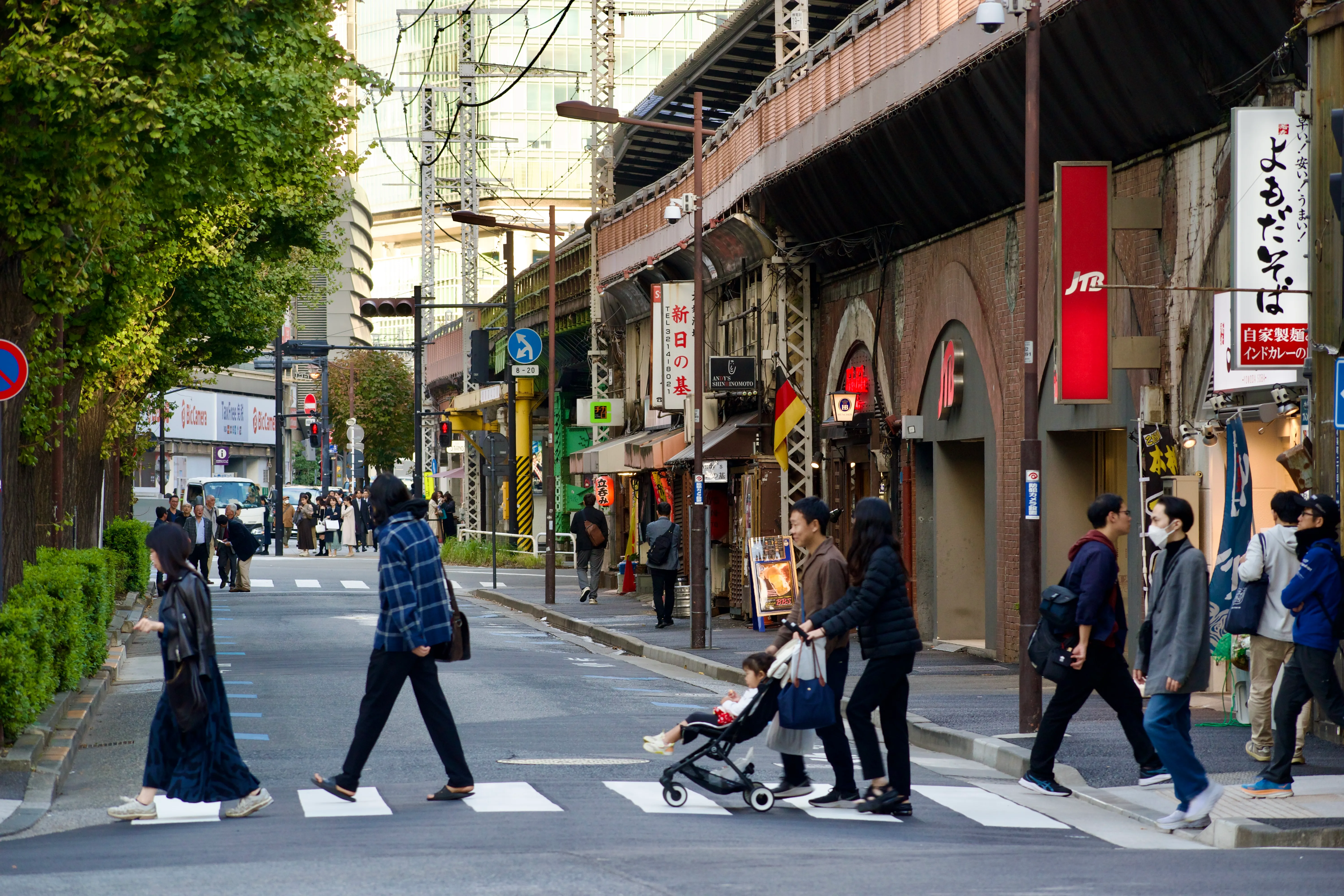 People cross a narrowish street in Tokyo in front of several shops built under train tracks