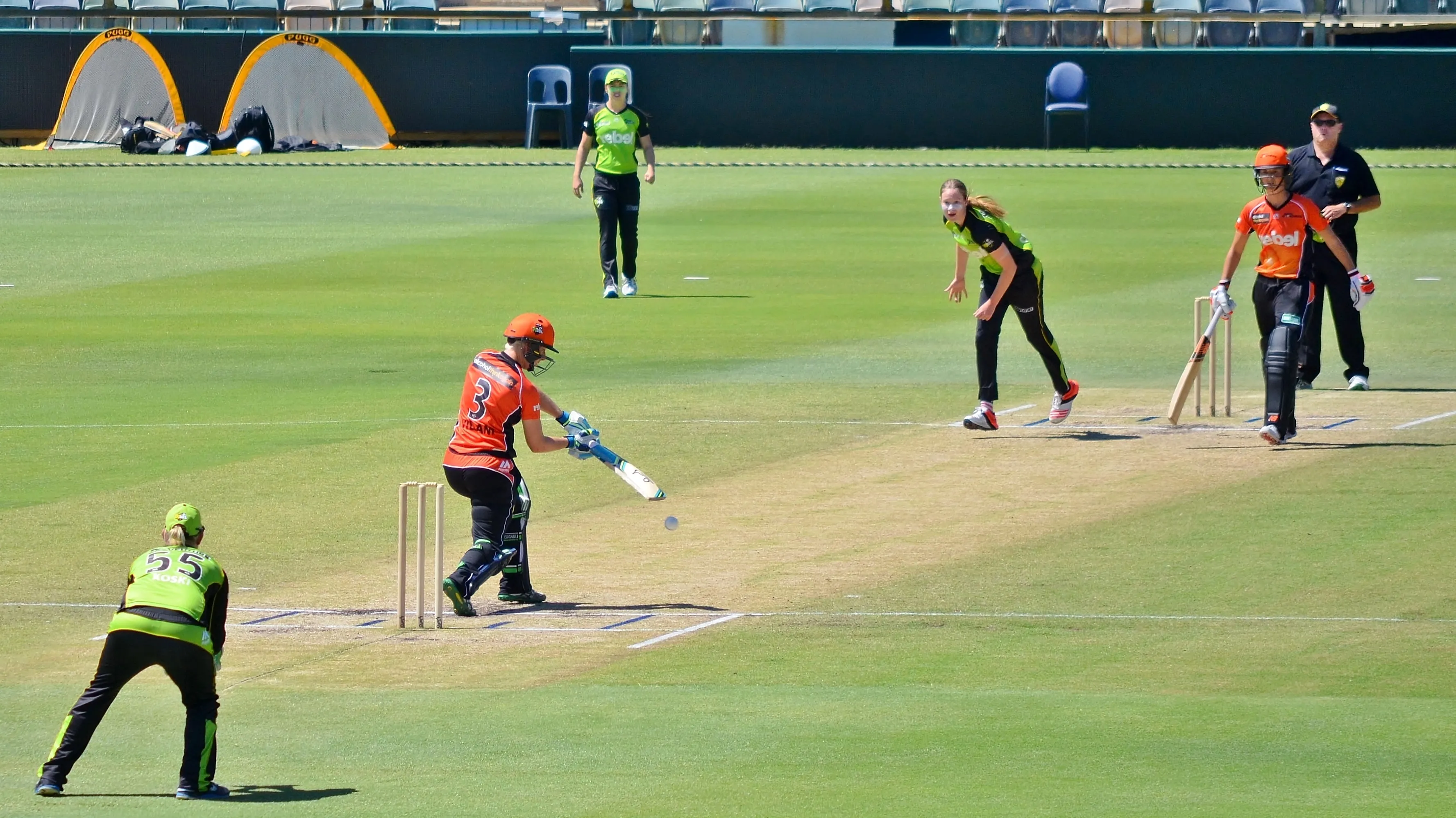 A batter strikes at the ball as the wicketkeeper, fielder, non-striker, and bowler react. (Bahnfrend / Creative Commons BY-SA)