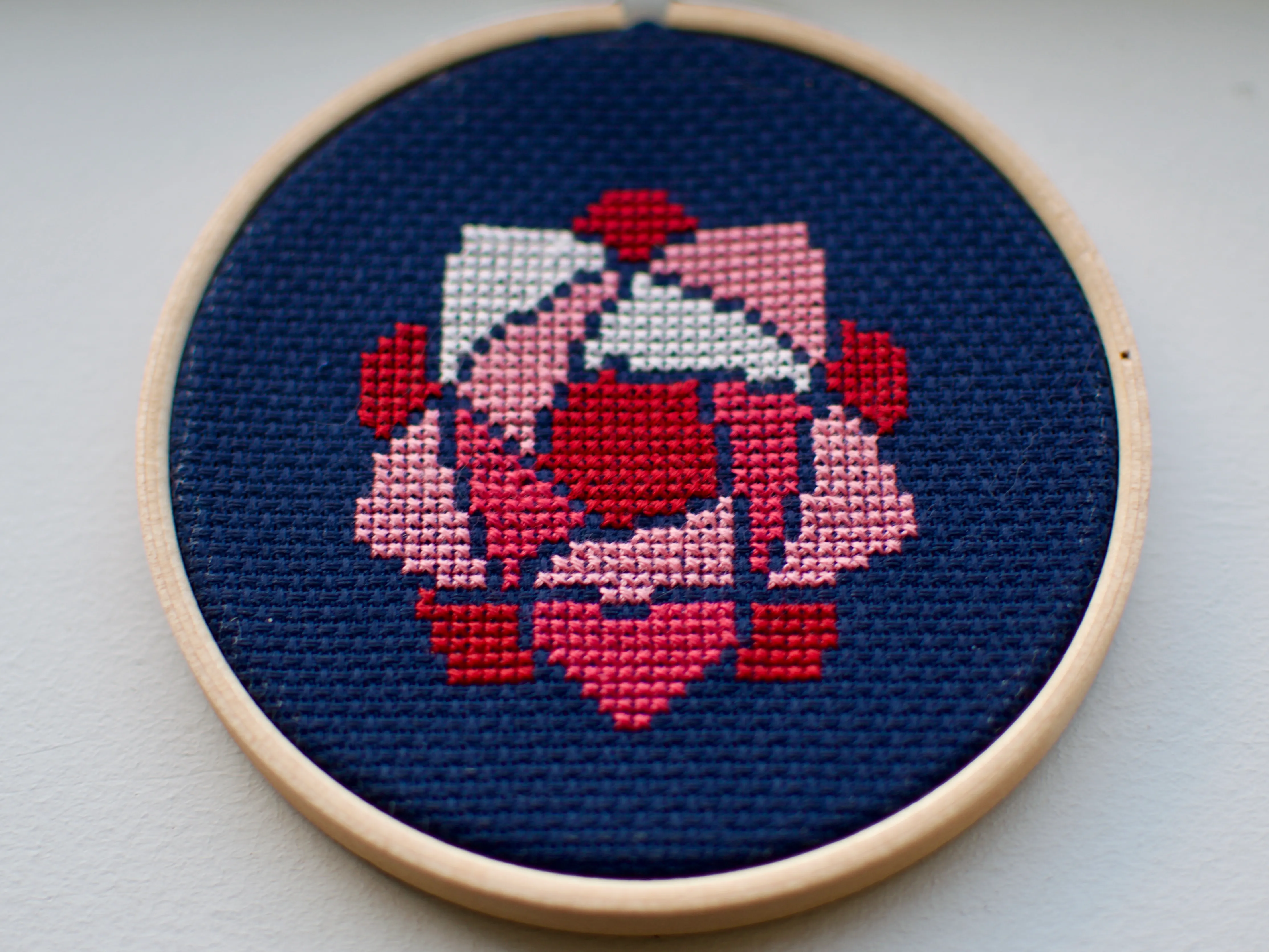 A small cross-stitch of a pink, roselike flower against a navy background