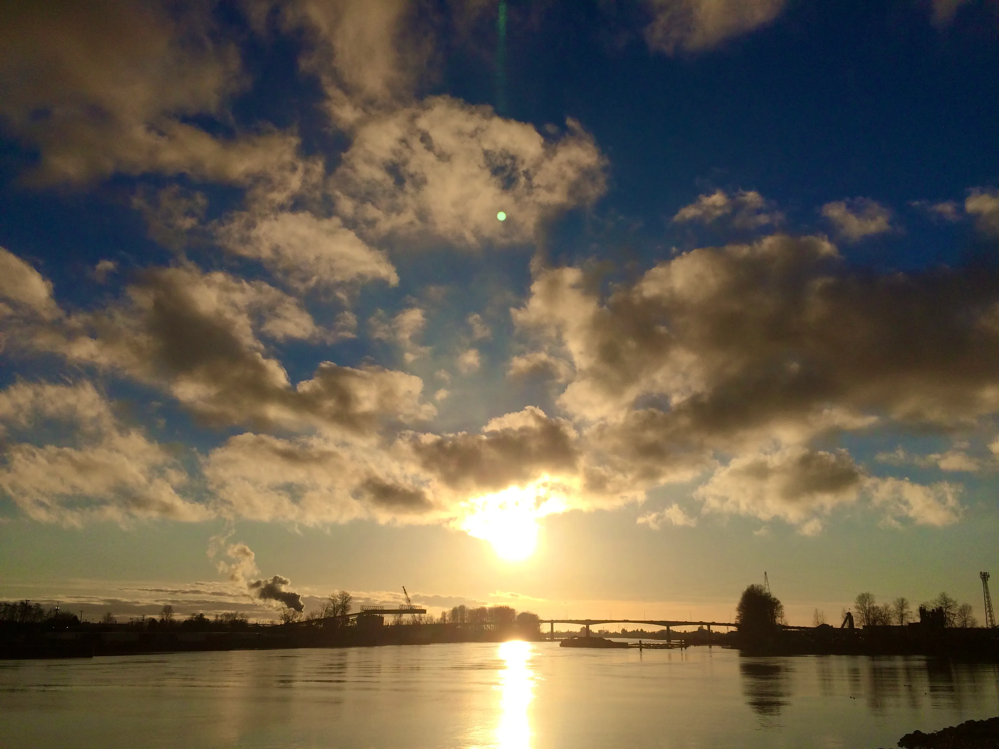 The sun nears the horizon over the Fraser River and the Knight Street bridge. The sky, dotted with clouds, fades from yellow on the horizon to deep blue overhead.