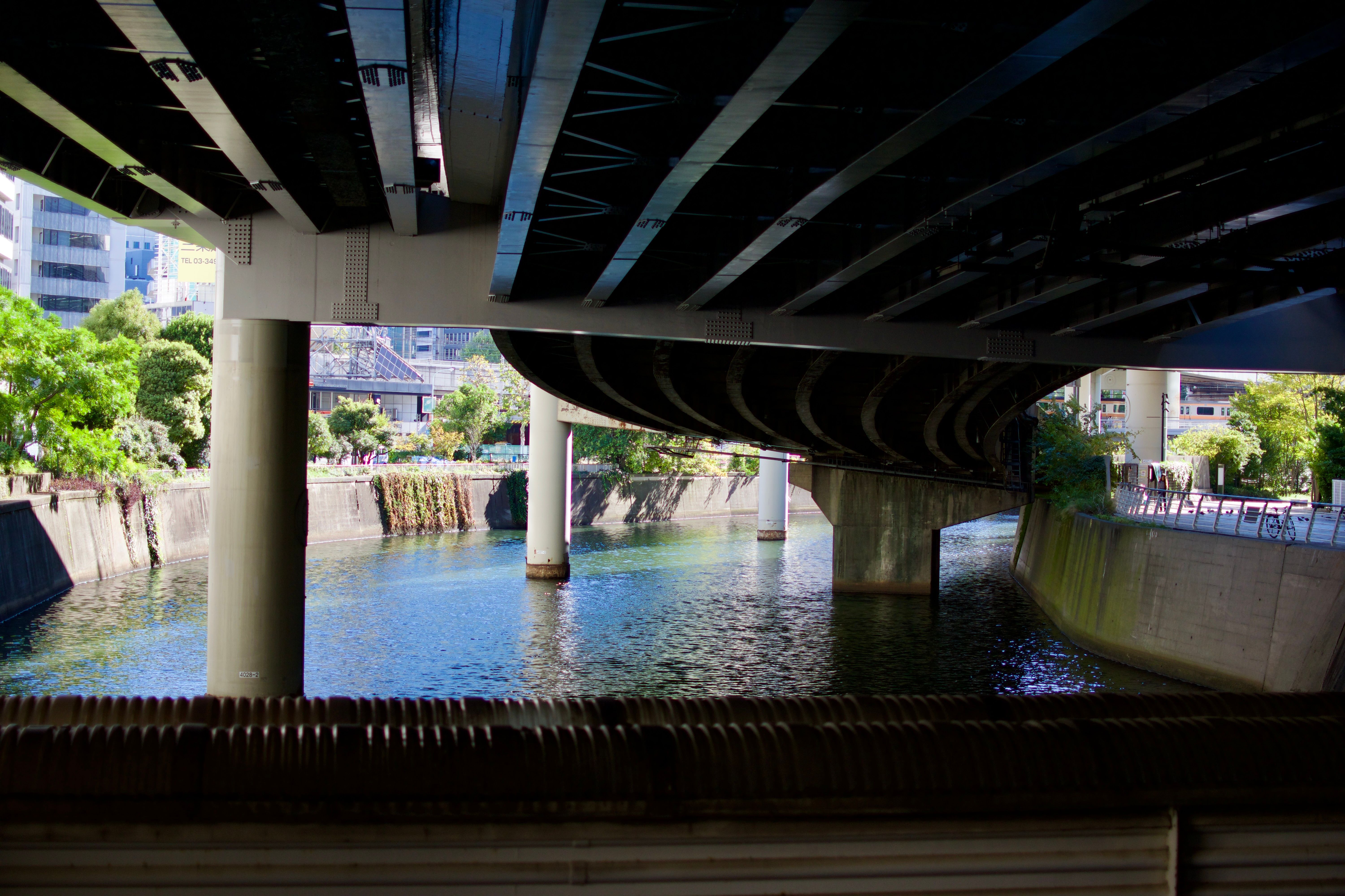 The underside of Tōkyō's metropolitan expressway, suspended above a river. The far side of the river is bathed in sunlight, contrasting with the shadows cast by the overpass.
