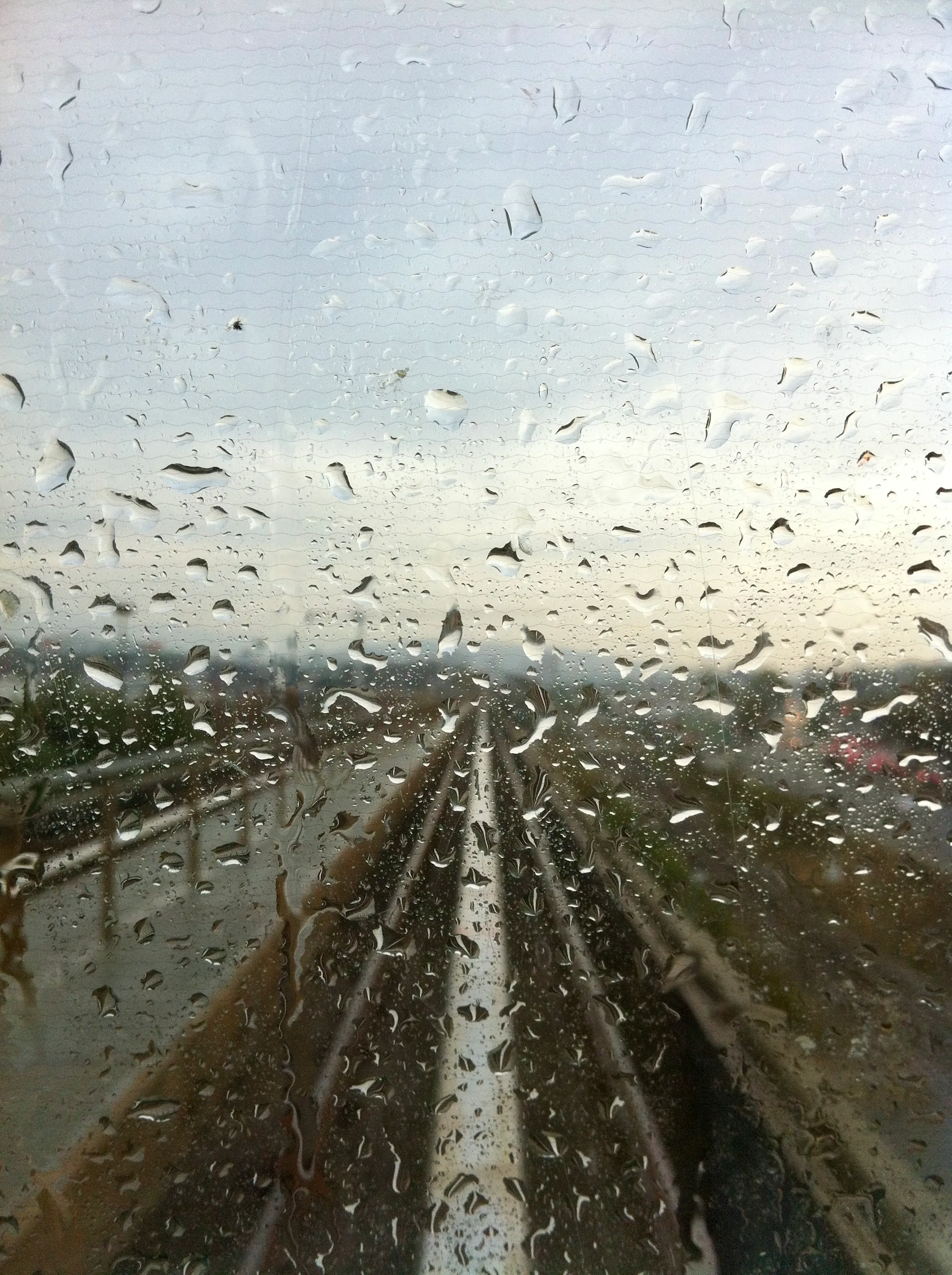 Raindrops streak down a window, obscuring a view of the train tracks ahead. The light suggests that the sun may be setting soon in clearer skies to the right of frame.