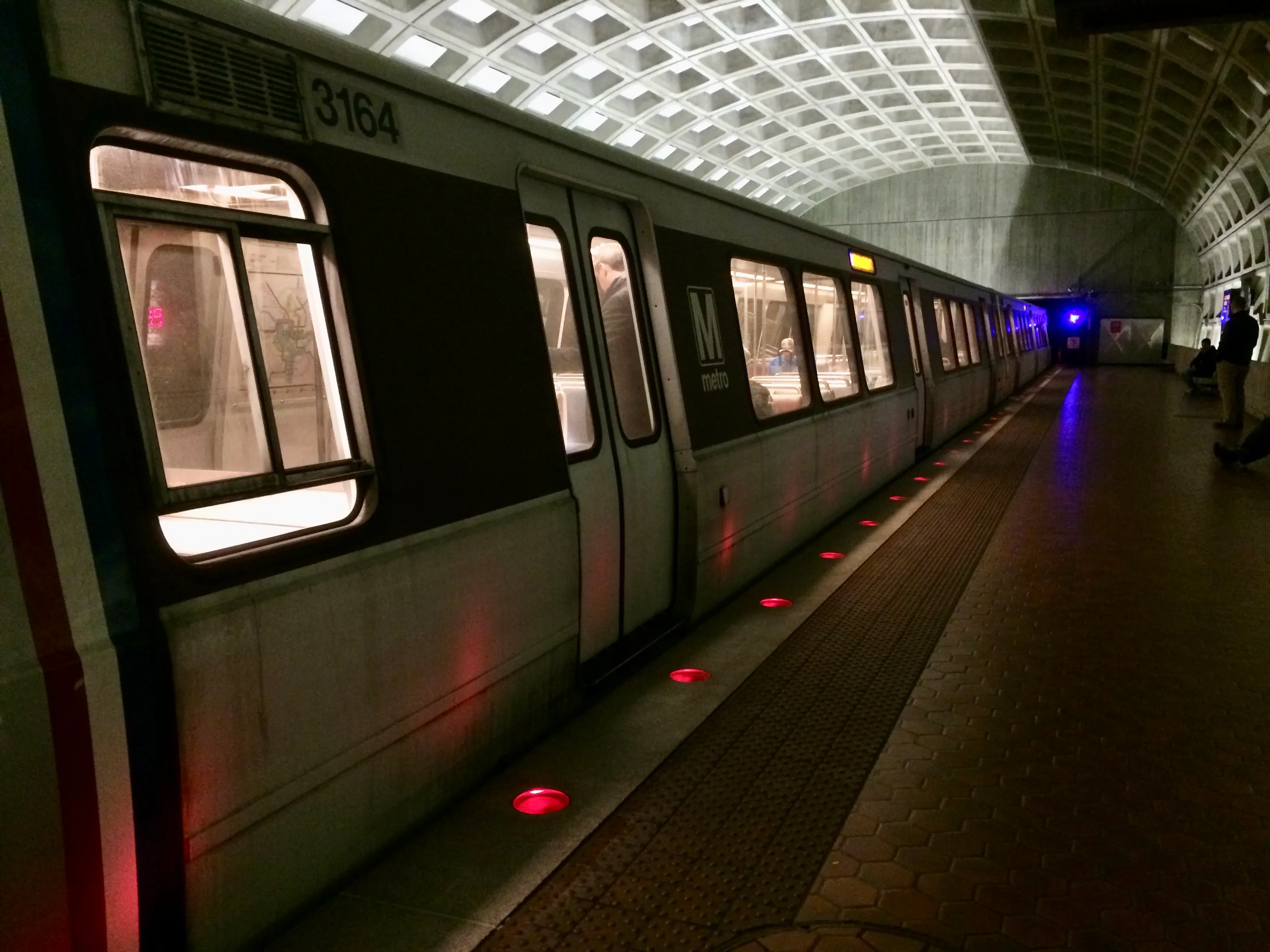 A subway train is stopped at a platform. Red lights mark the edge of the platform, and the elliptical ceiling is half-illuminated.