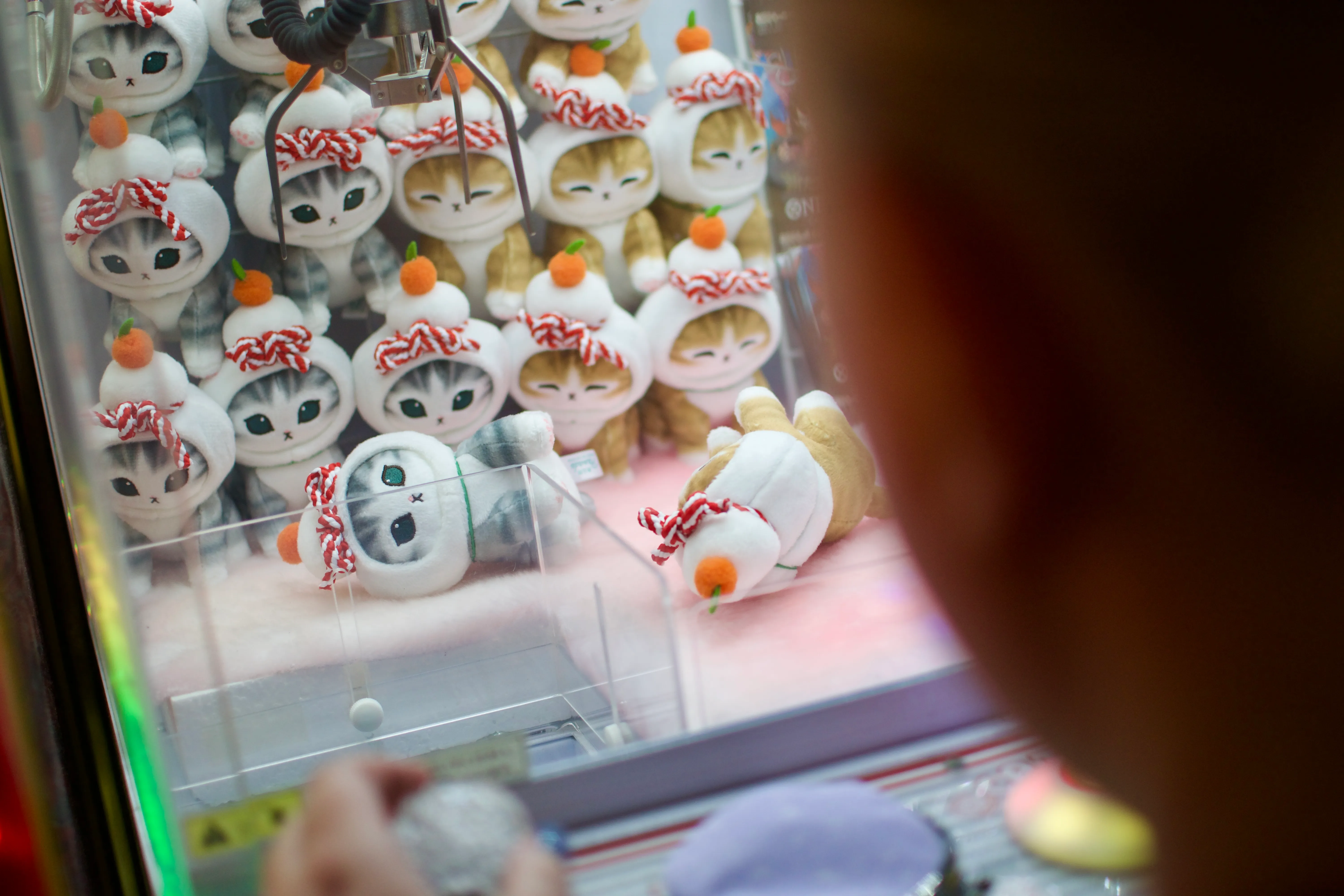 An over-the-shoulder action shot of a crane game, with the claw hovering over a plush of a cat wearing a hat with a mandarin orange on it