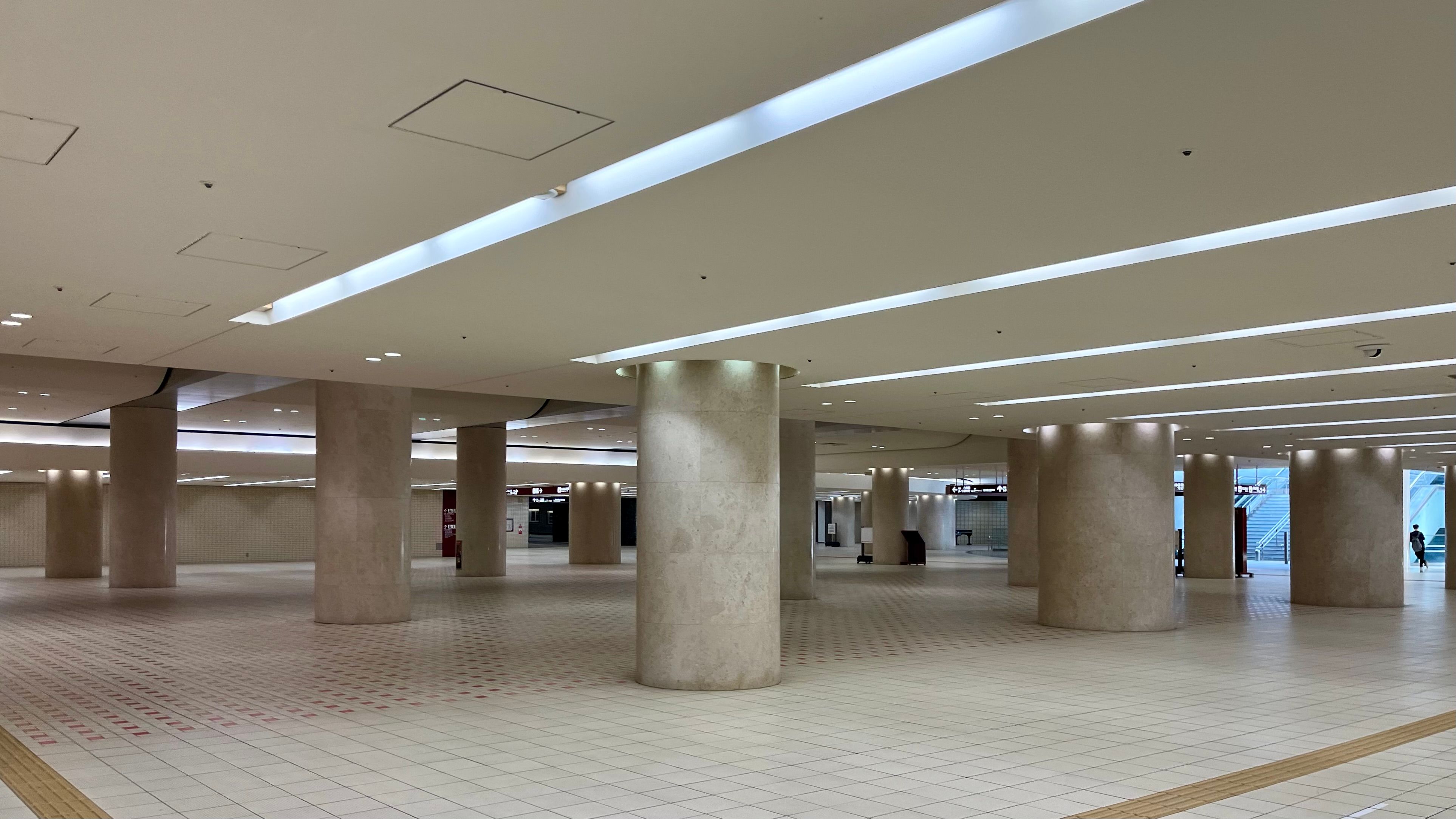 A large room with a low ceiling supported by many wide concrete pillars. The room has tiled white floors, fluorescent lighting, and is completely empty aside from the rows of pillars.