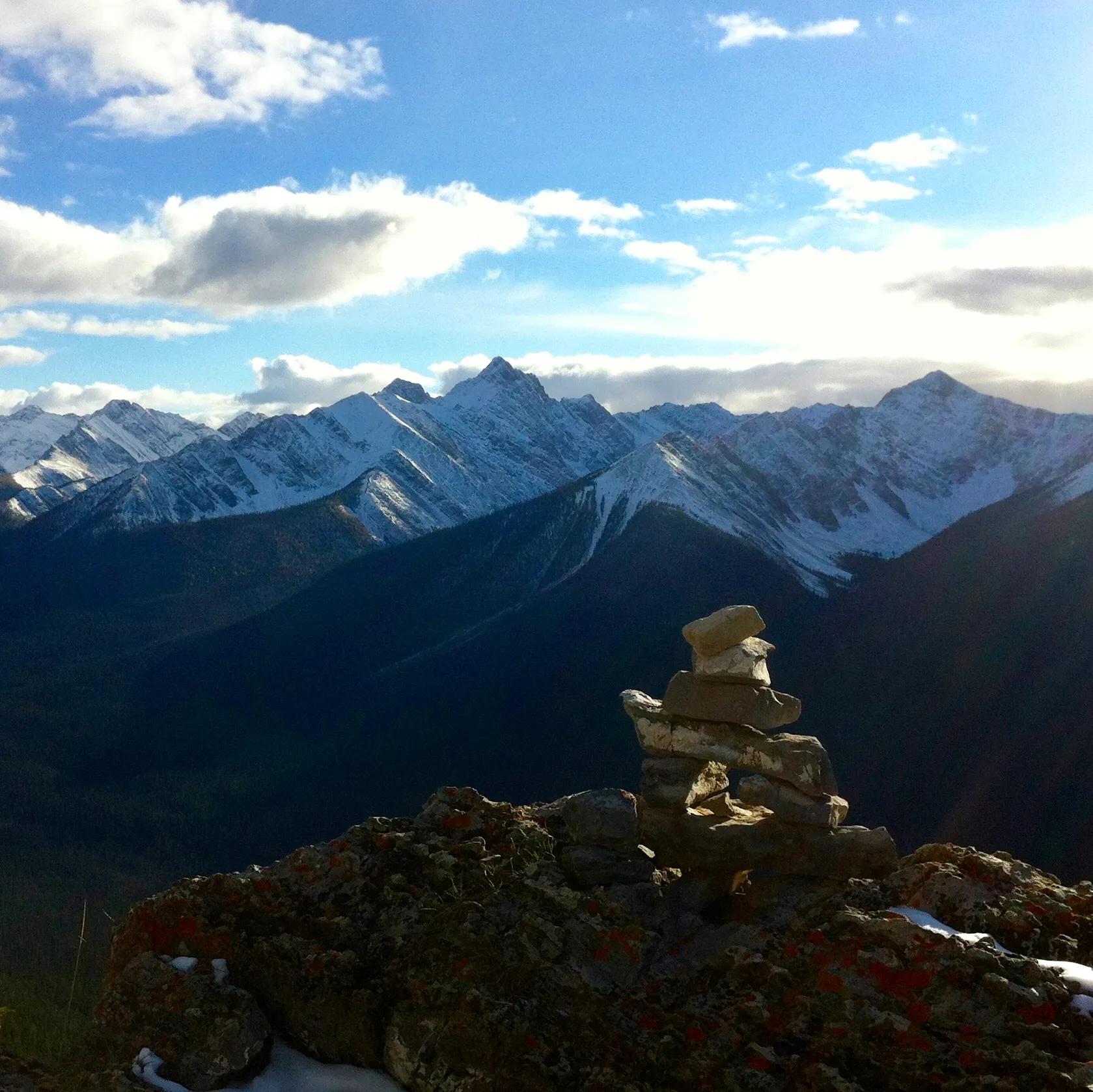 An inukshuk on a rocky outcrop with a backdrop of snow-covered mountains under a cloudy sky.