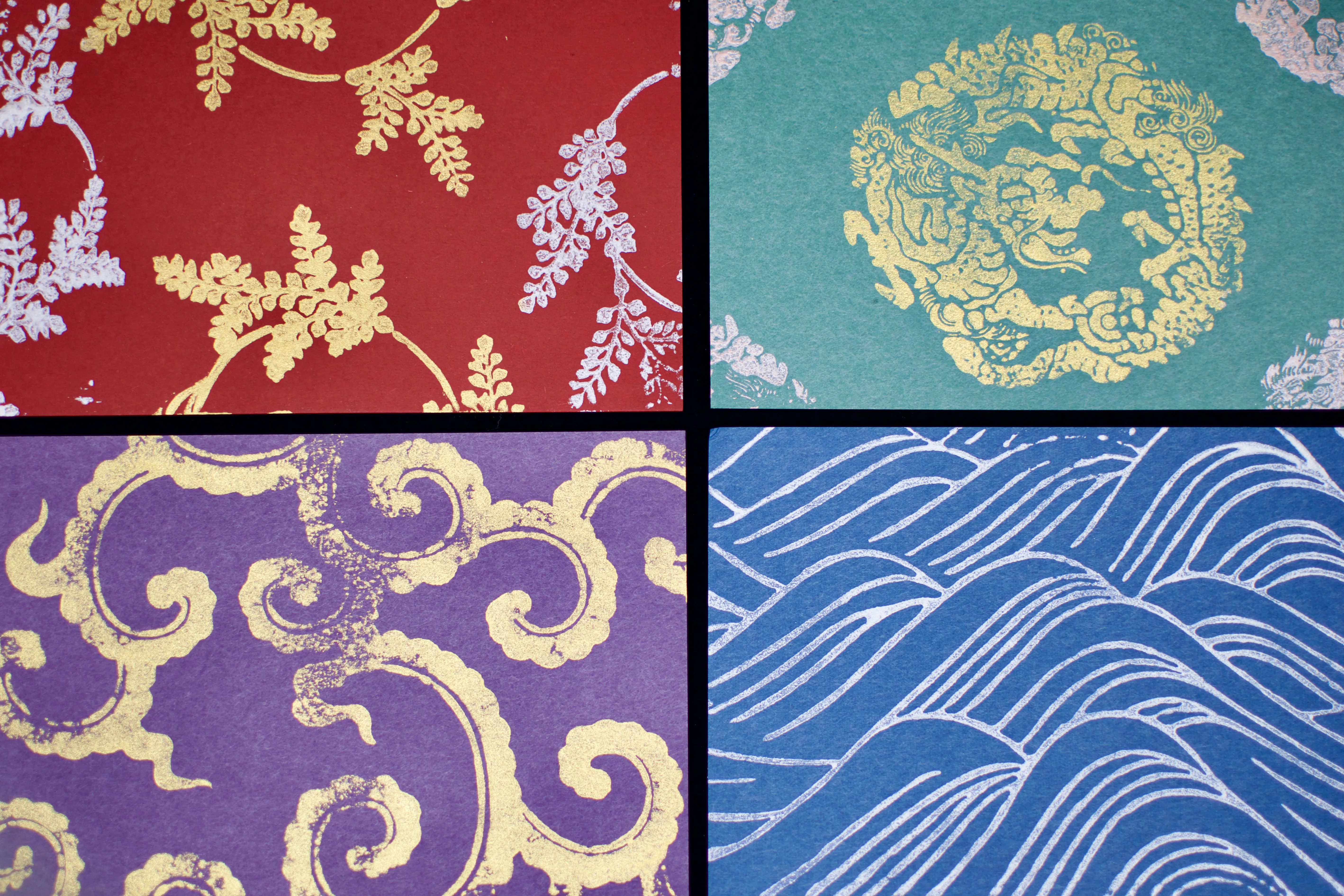 Four postcard-sized woodblock prints: silver and gold ferns on red paper; a gold dragon on green paper; gold clouds on purple paper, and silver waves on blue paper