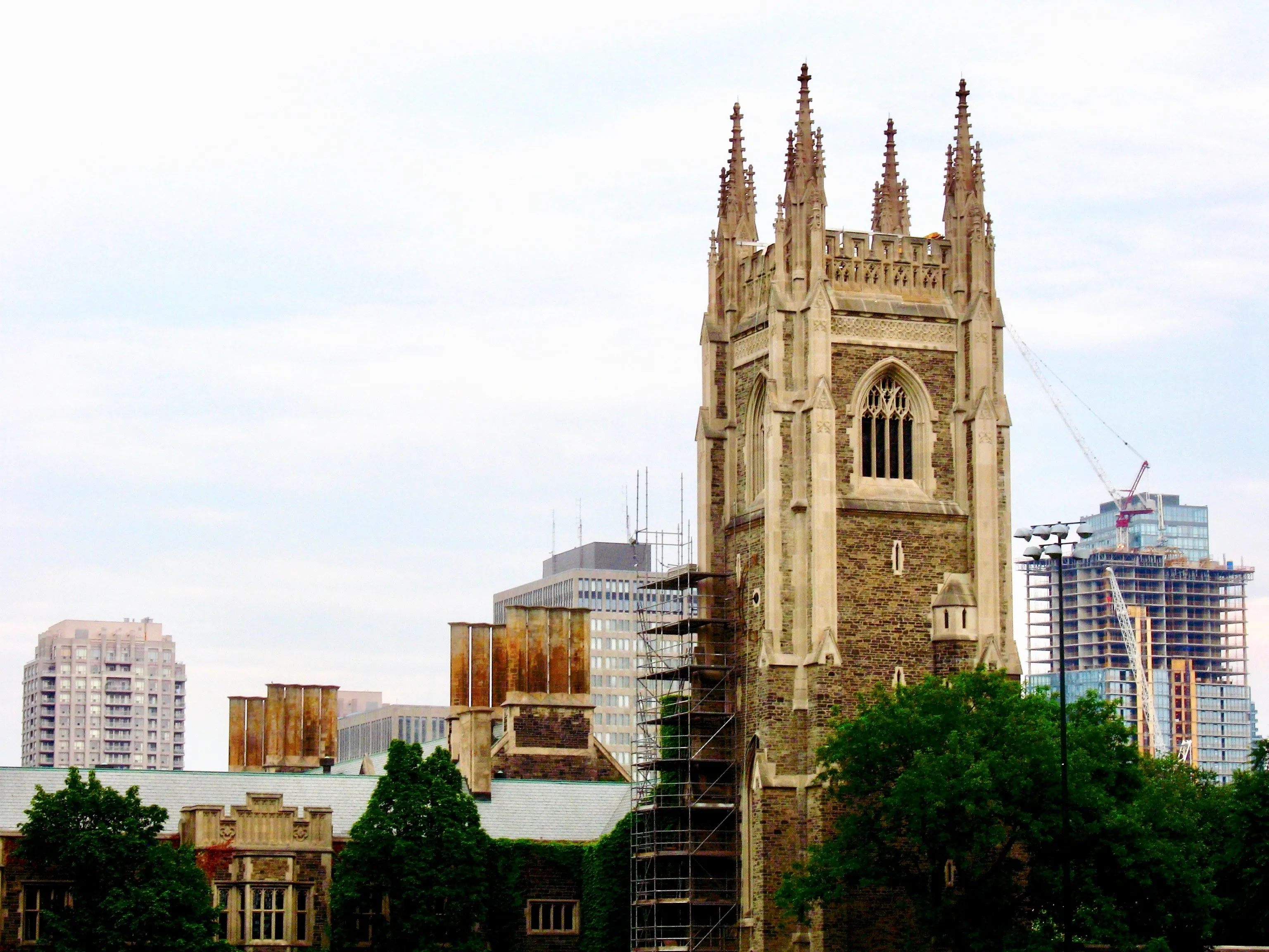 A stone church tower is being repaired with scaffolding, while a modern glass skyscraper is being constructed in the background.