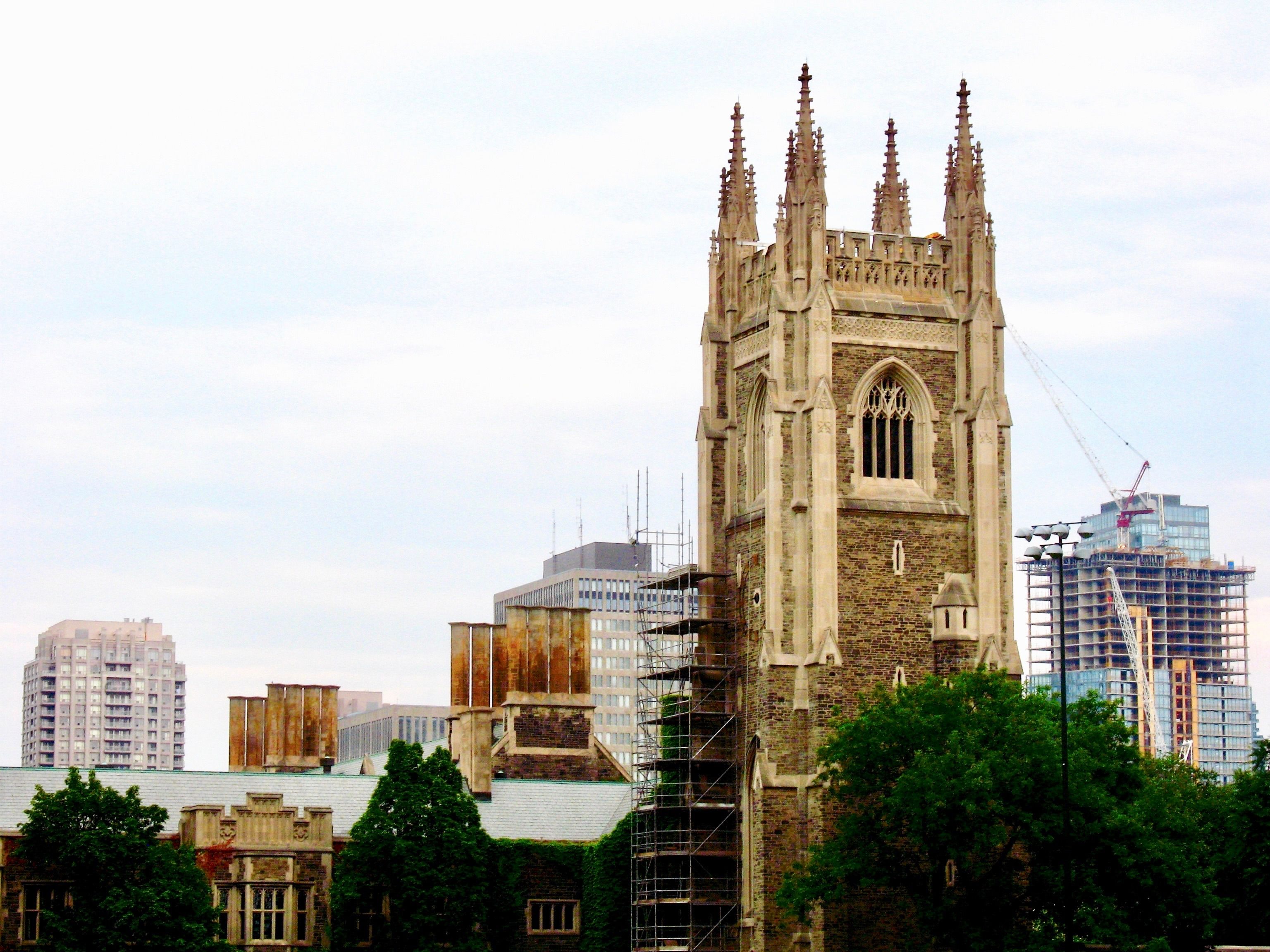 A stone church tower is being repaired with scaffolding, while a modern glass skyscraper is being constructed in the background.