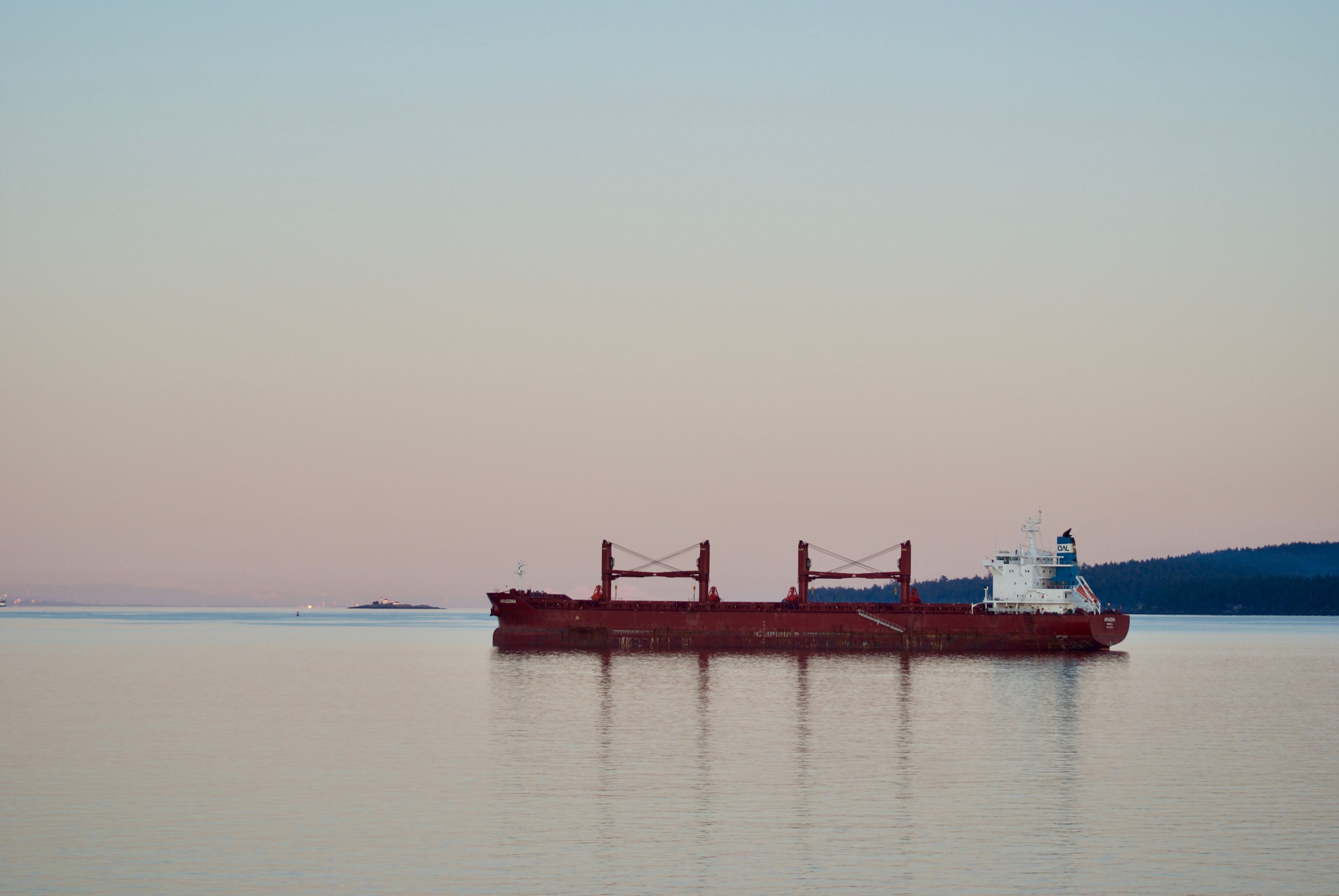 An empty red cargo ship is moored at sea. The clear sky has the pastel colours of sunset, transitioning from red at the horizon to greenish-blue at the top of the frame. The cranes on the cargo ship are reflected on the calm waters.