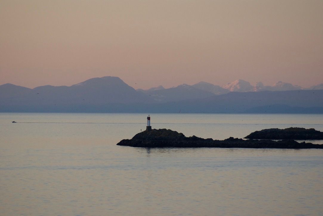 A starboard-hand daybeacon marks a rocky outcropping on the sea at sunset. The wake of a small boat traces a straight line across the still water. Purple mountains and a hazy, pink-orange sunset are in the background.