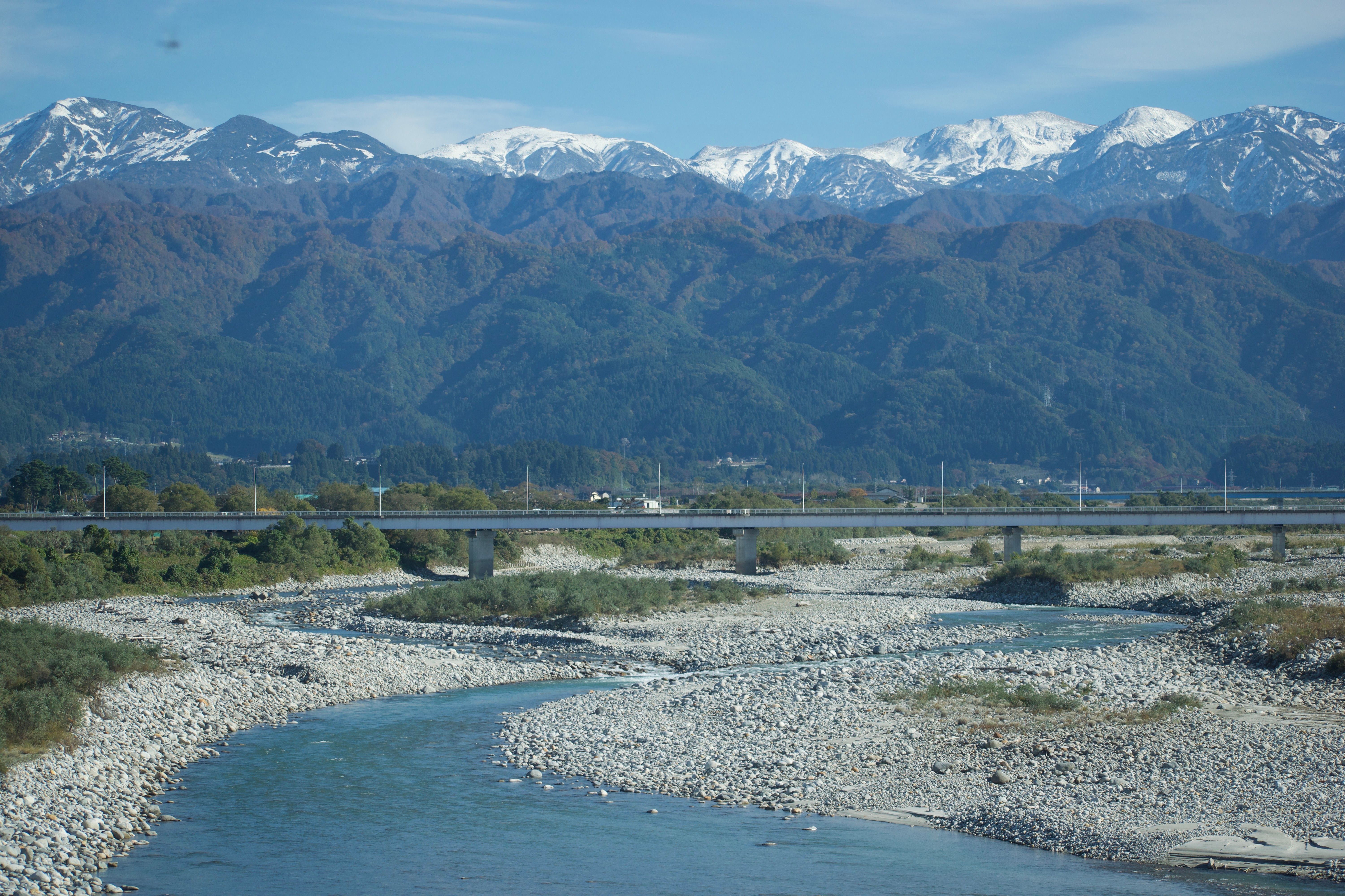 A wide river flows between gravel banks, spanned by a concrete bridge. Snow-capped mountains rise steeply in the background.