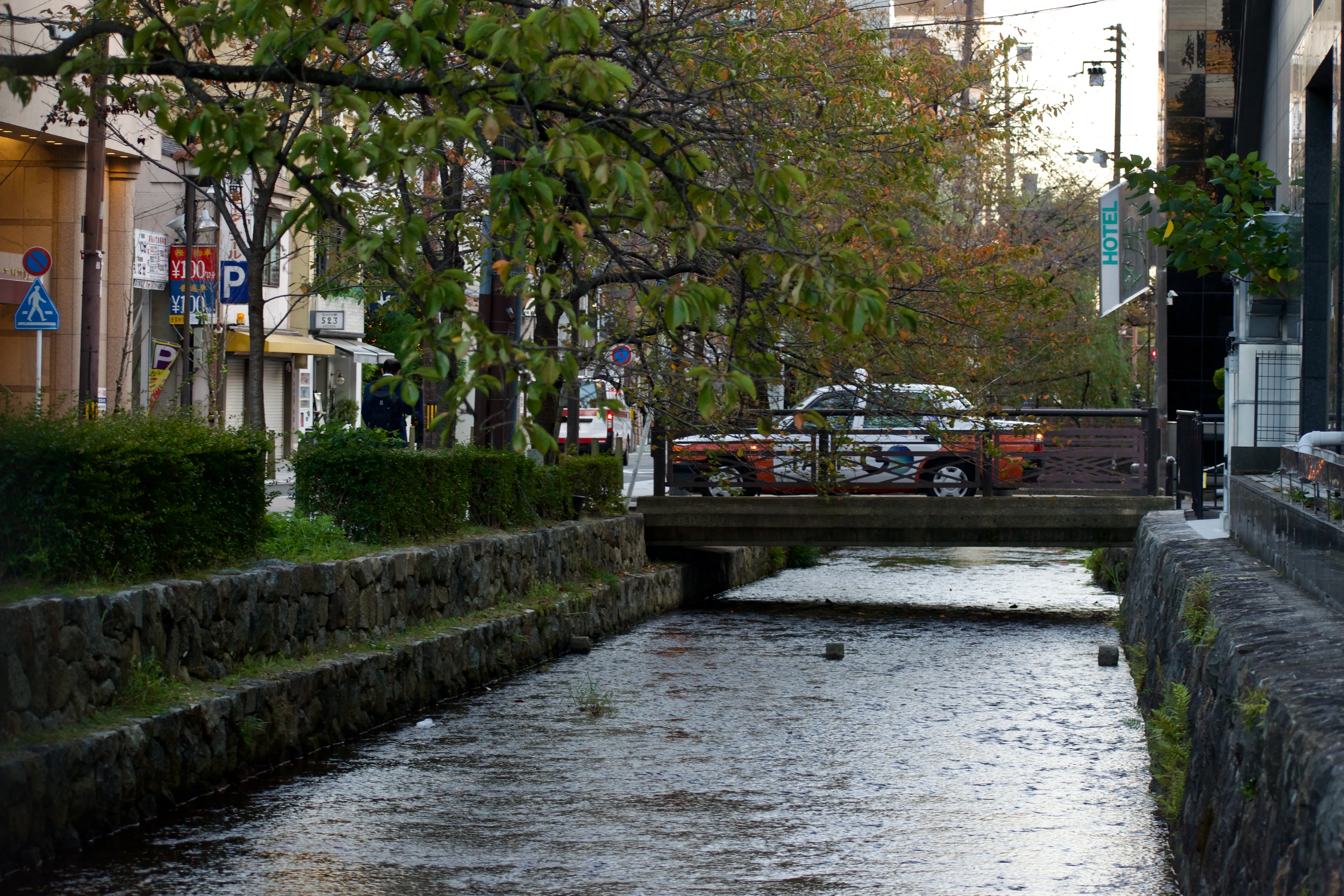 A taxi waits on a tiny bridge, barely longer than the taxi itself, which spans a narrow, shallow canal flanked by trees.