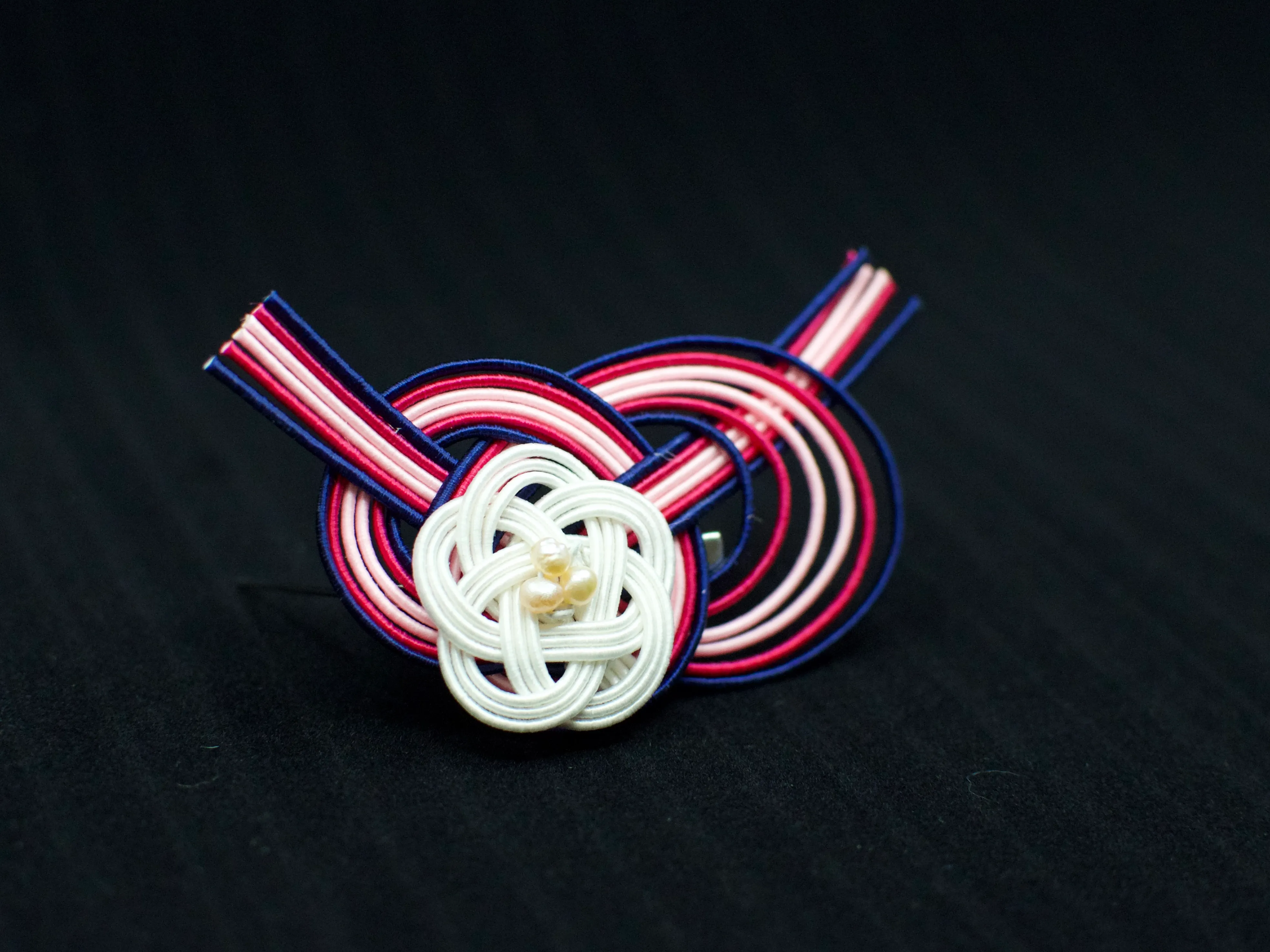 A brooch in the shape of a knot, made from navy, fuchsia, and pink paper cords. A smaller white knot with pearls is on top.