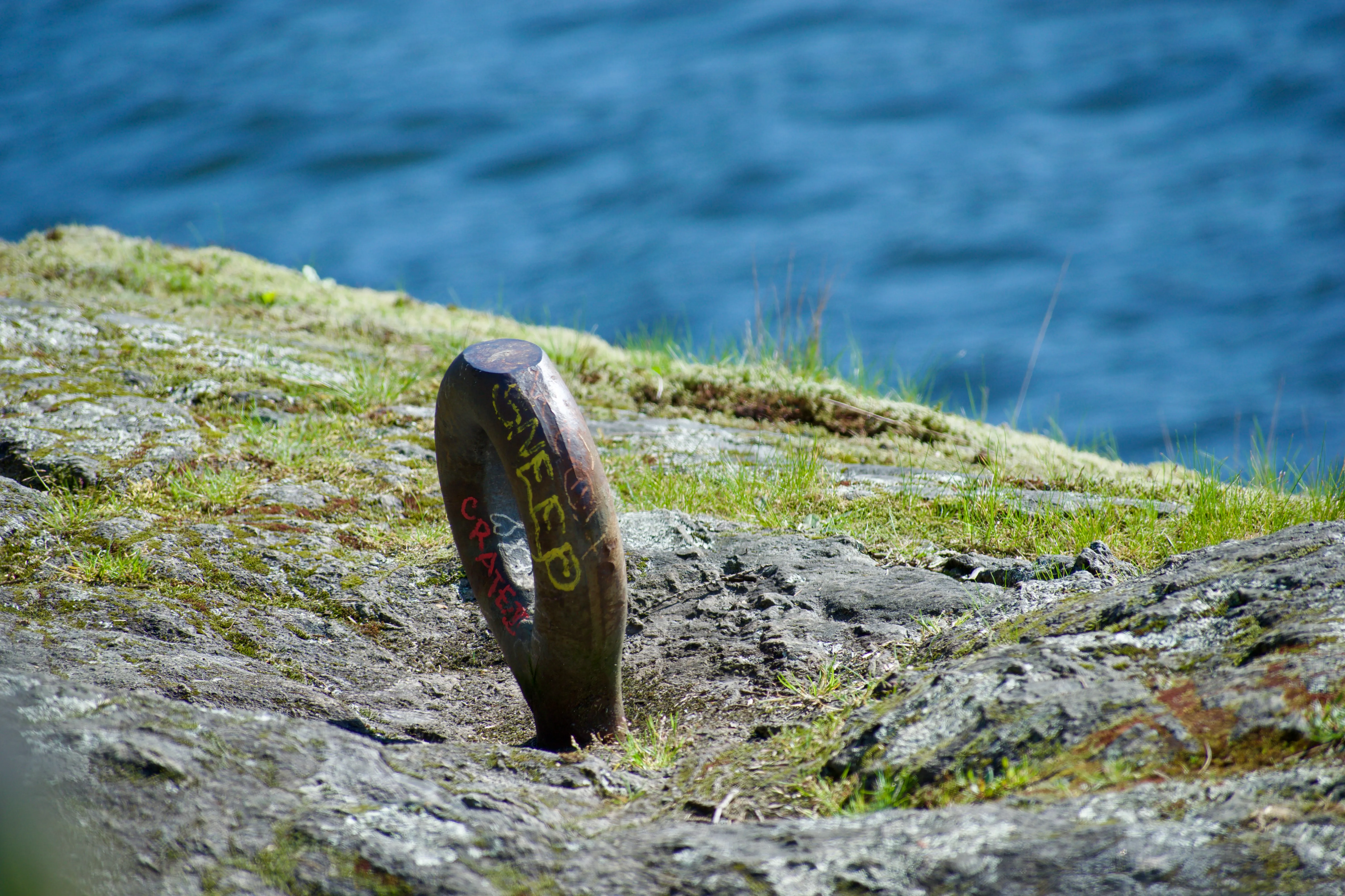 A metal post is driven into a mossy rock with the blue water of Burrard Inlet in the background.