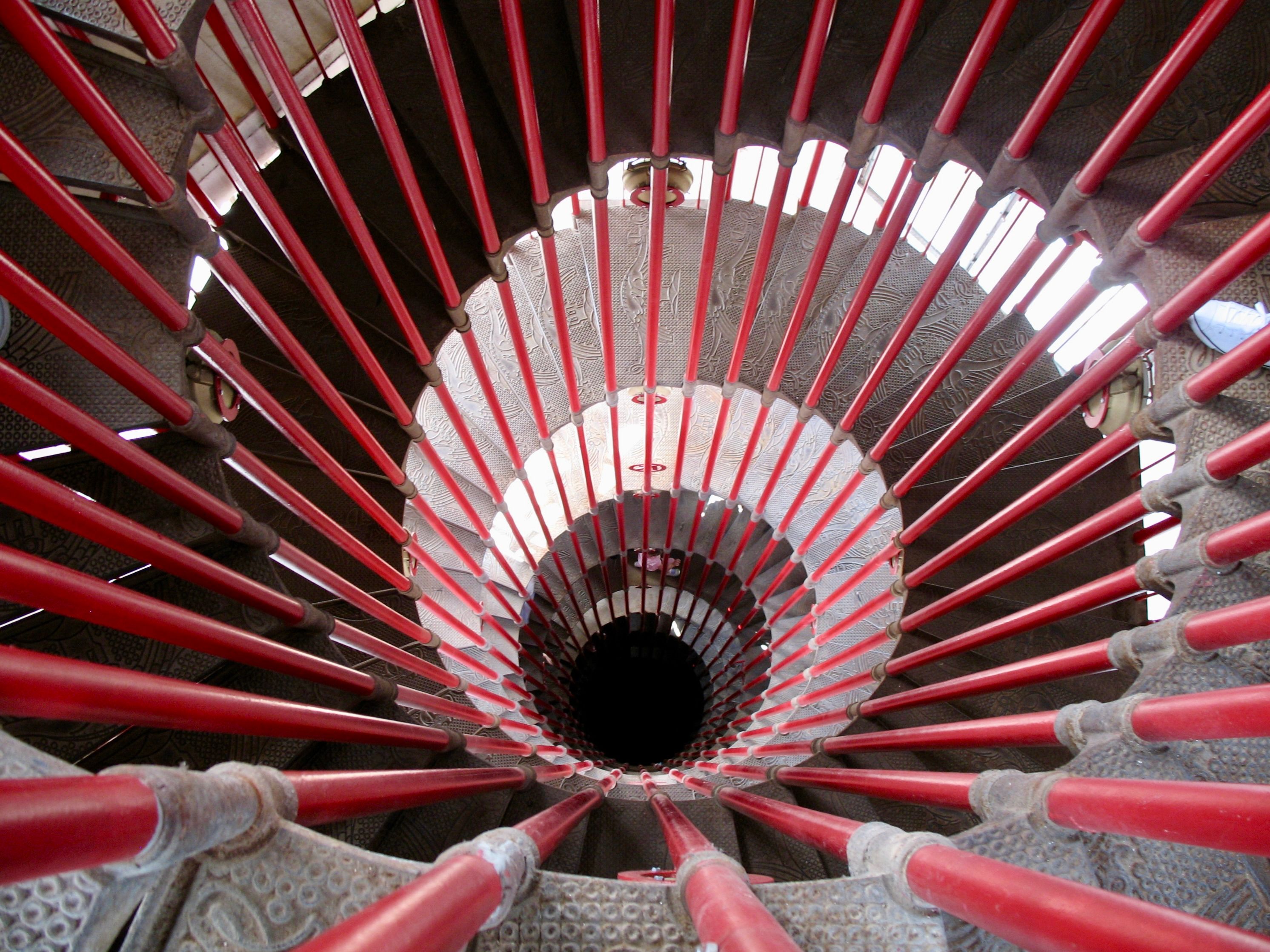 A view down the center of a narrow double-helical spiral staircase several stories high. The focus is on the red metal beams enclosing the inner circle, emanating from the center of the frame outwards.