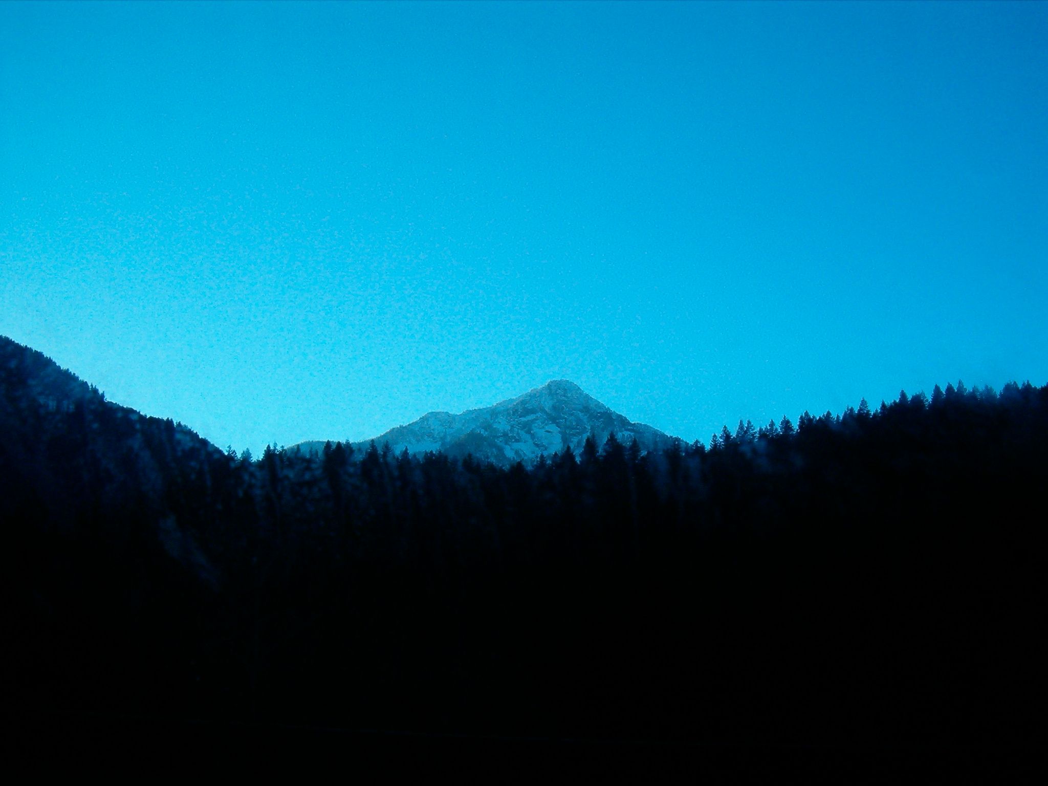 Snow-covered mountain peak rises prominently above a dark, forested landscape. The peak is sharply defined against a dusky sky.
