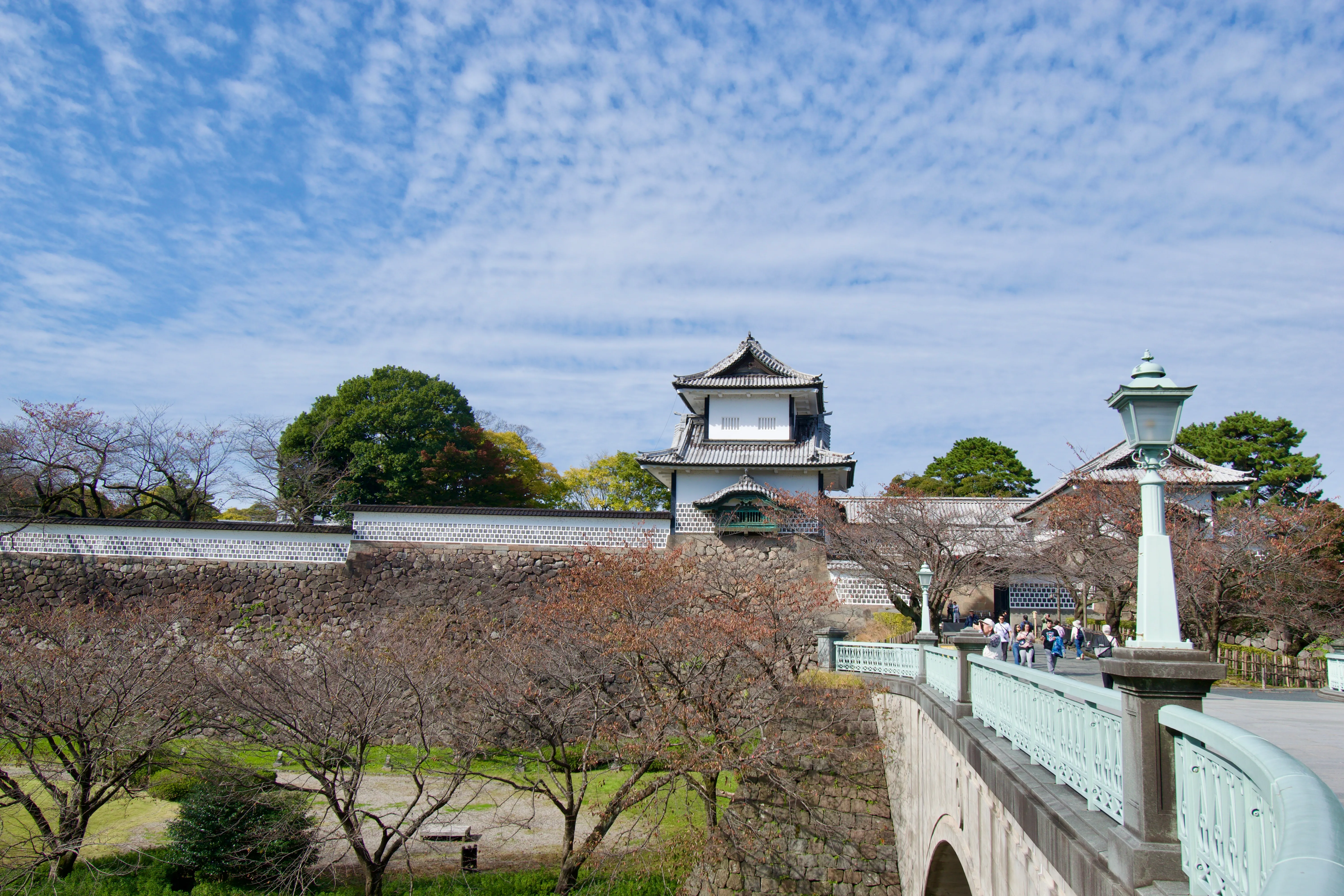 A stone bridge leads to the outer walls and turret gate of Kanazawa Castle. Trees line the street below the bridge, and the clear sky is dotted with clouds.