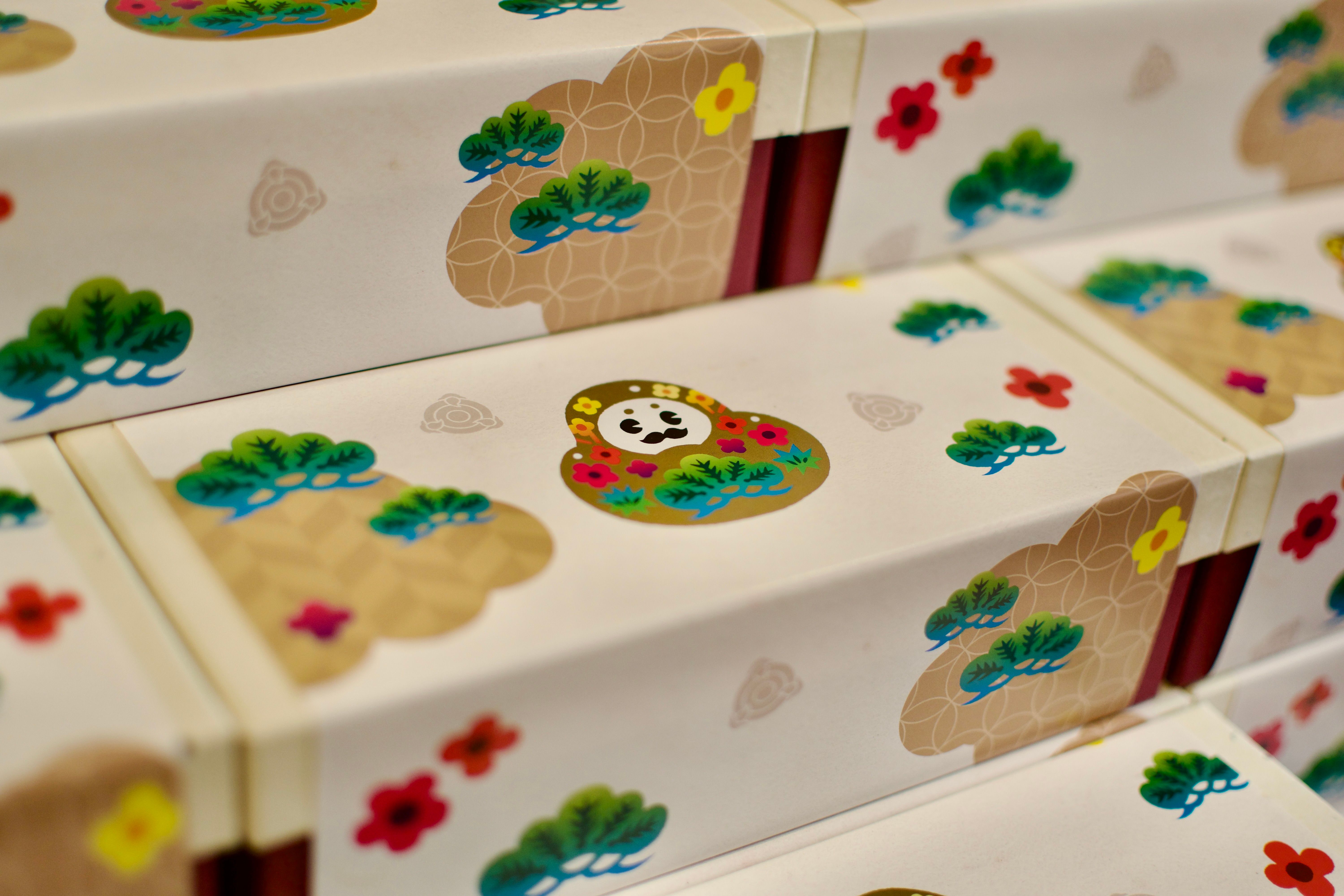 A box whose wrapping depicts a mustachioed round doll decorated with gold leaf, flowers, and trees.