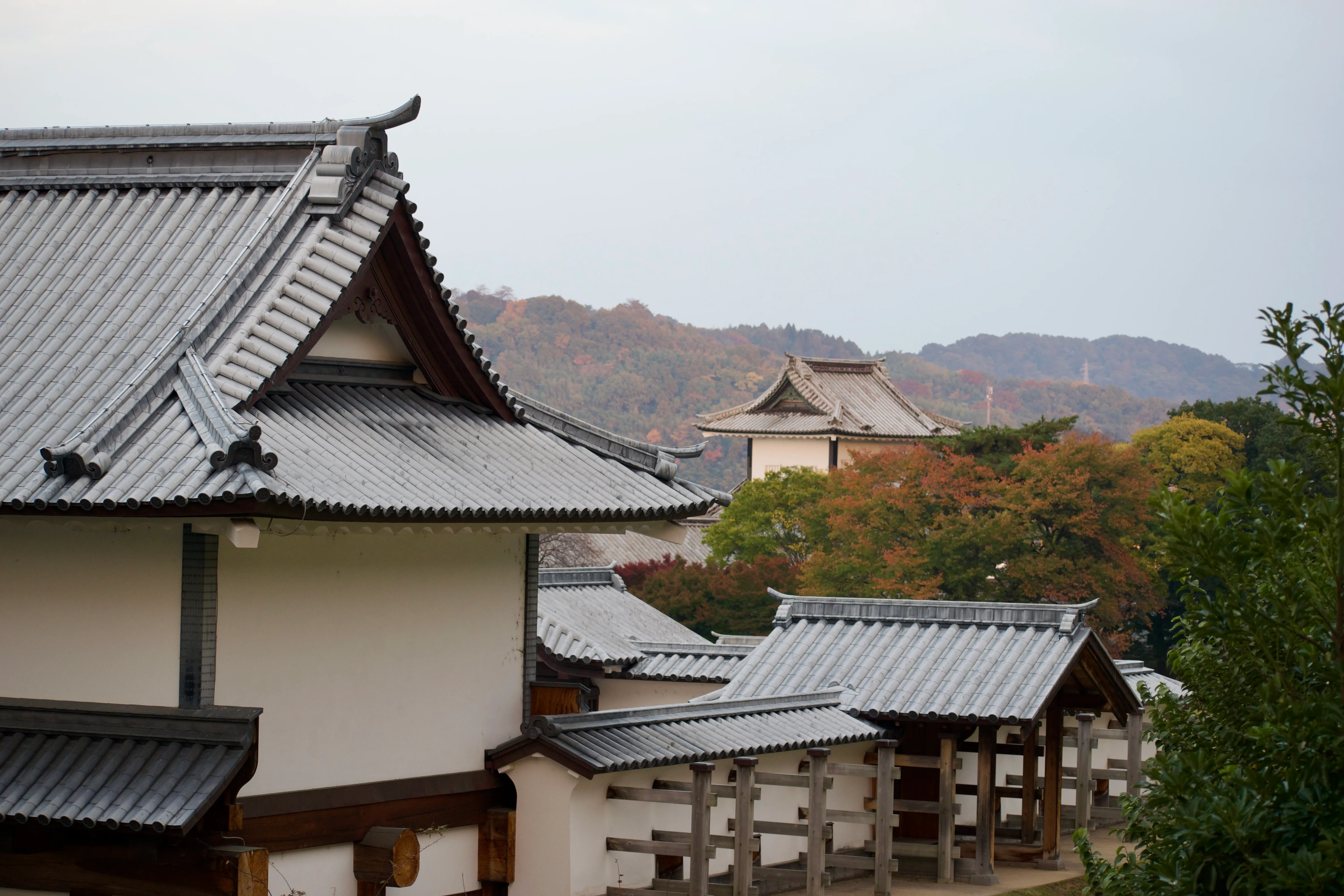 A back view of Kanazawa Castle's keep. In the background, autumn-coloured trees, a castle turret, and the forested hills of Utatsuyama are visible.