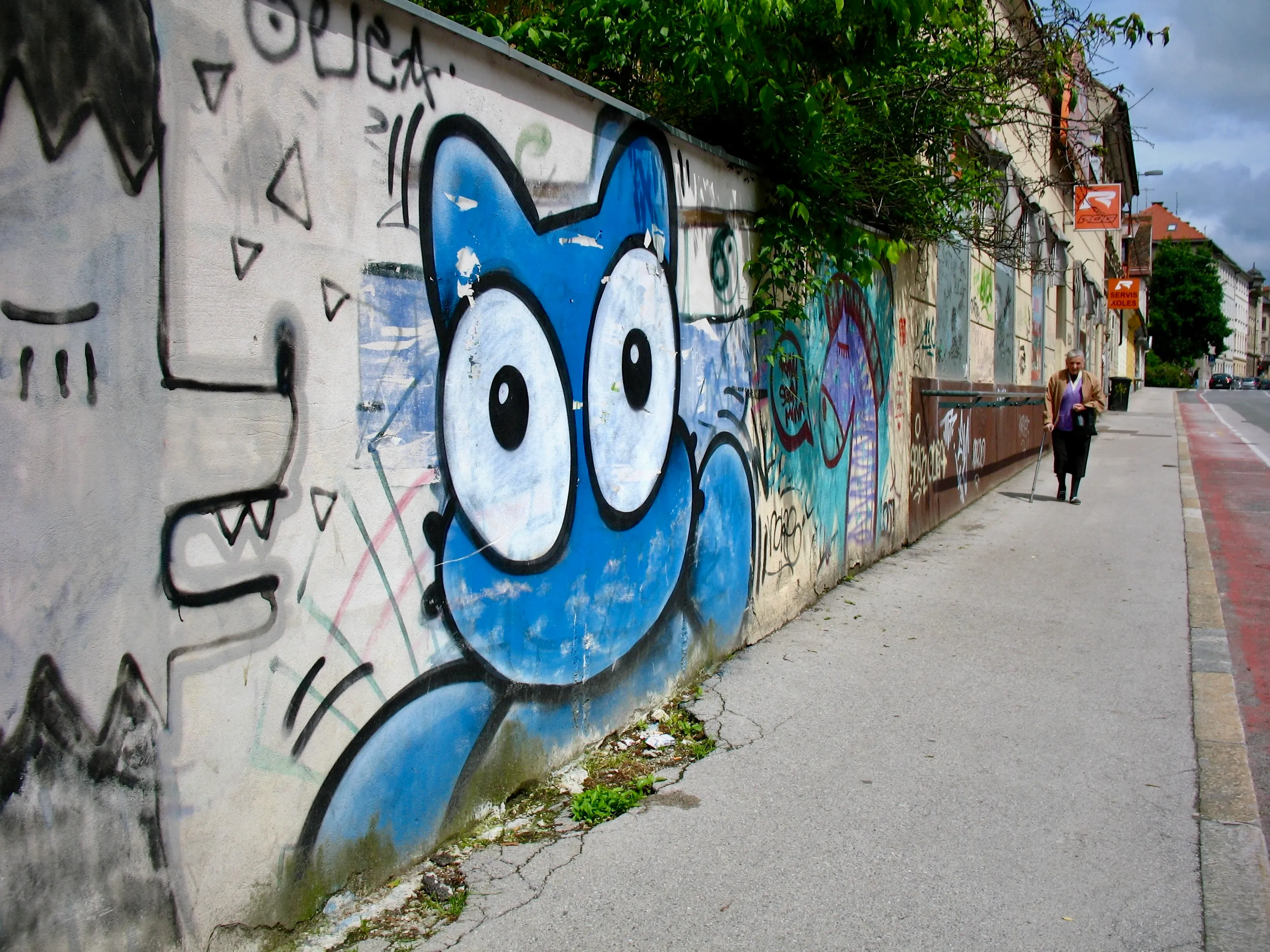A wall covered in large graffiti illustrations, including a blue cartoonish Moomin-like character. An elderly woman is walking with a cane on the adjoining sidewalk.