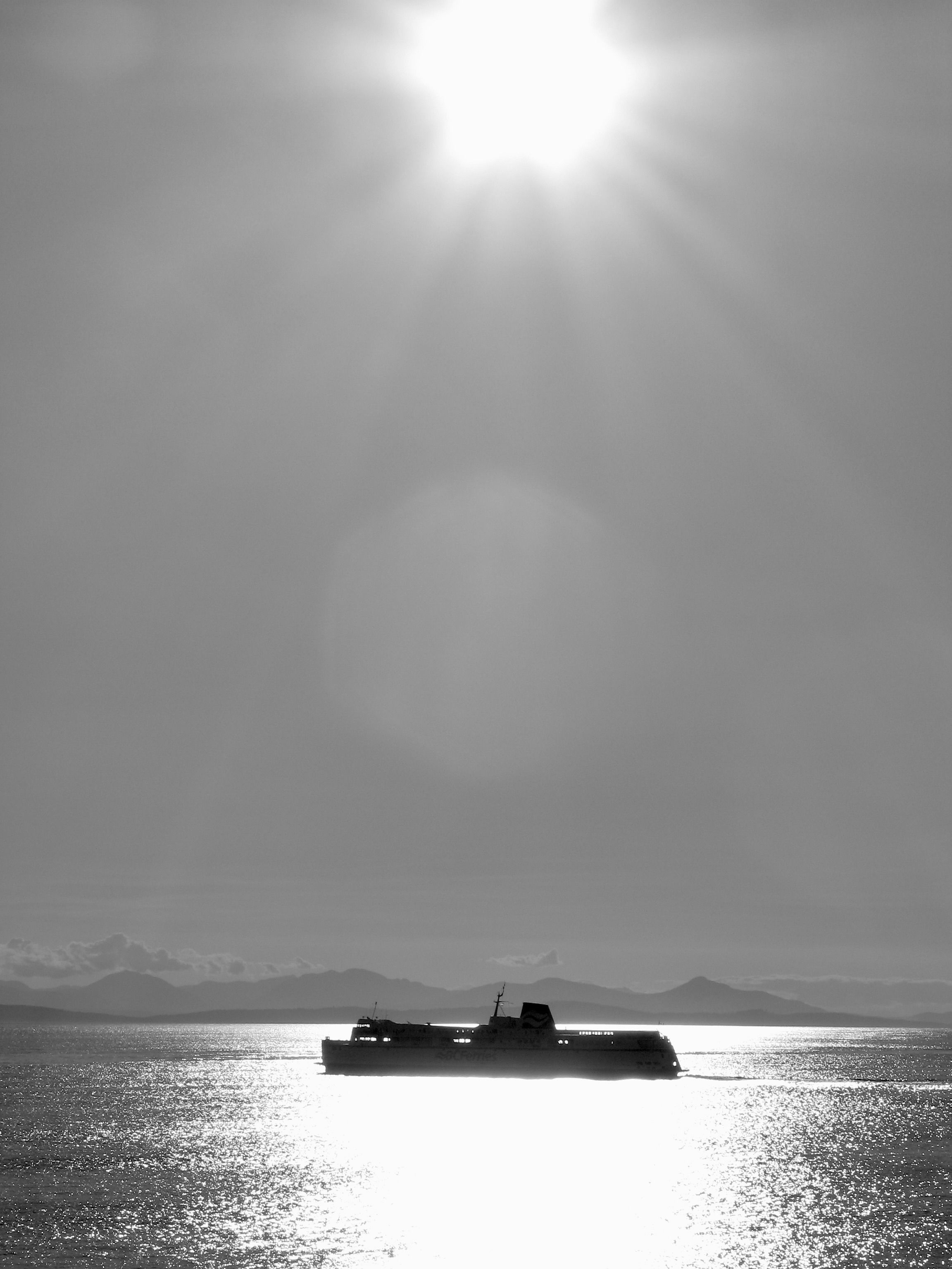 A black-and-white photo showing the silhouette of a ferry in the glare of the sun on the water.