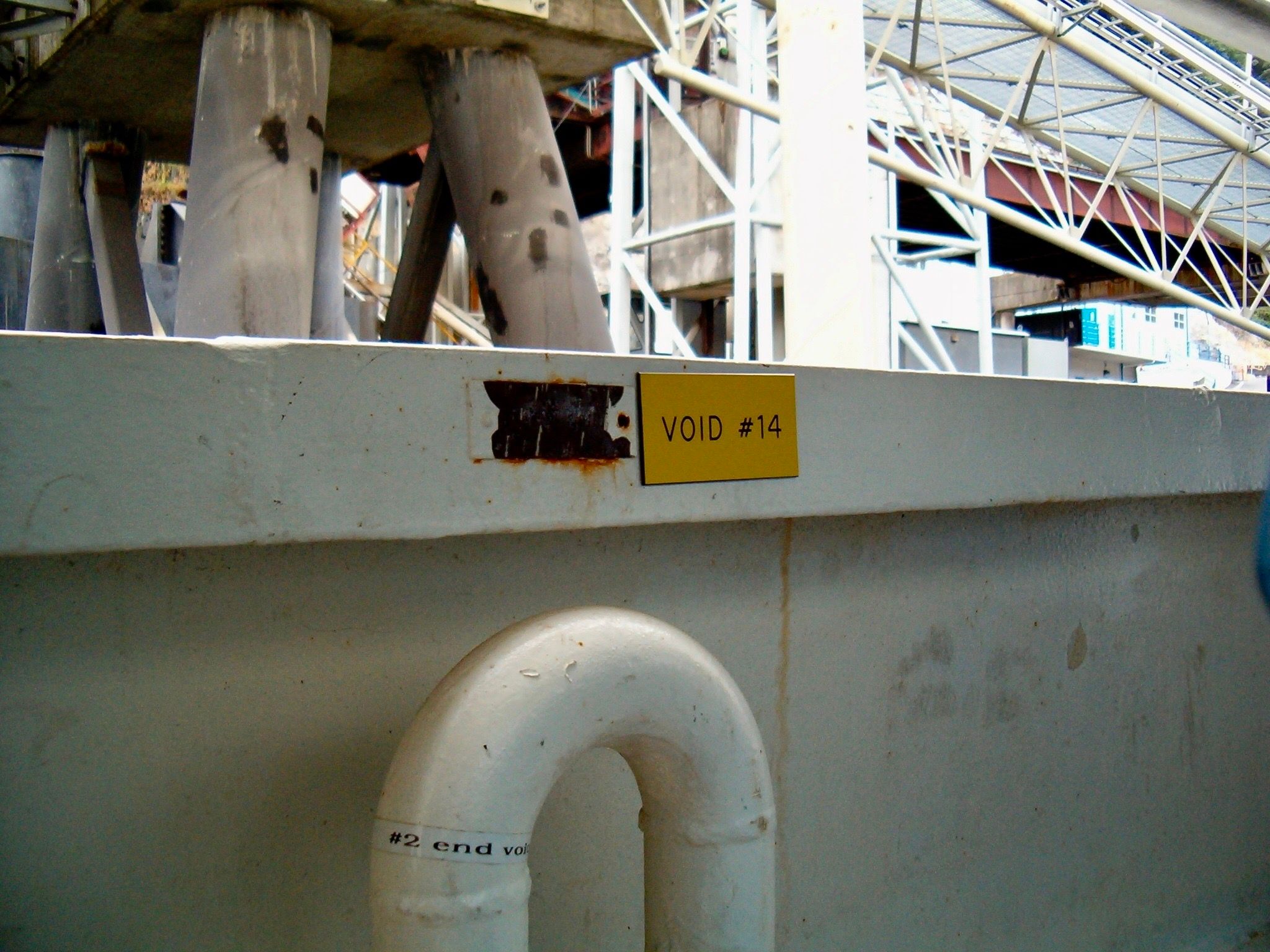 A yellow sign reading 'Void #14' labels the top edge of a stomach-high metal wall. Through the void can be seen concrete pillars and metal structures making up the dock infrastructure of a ferry terminal.