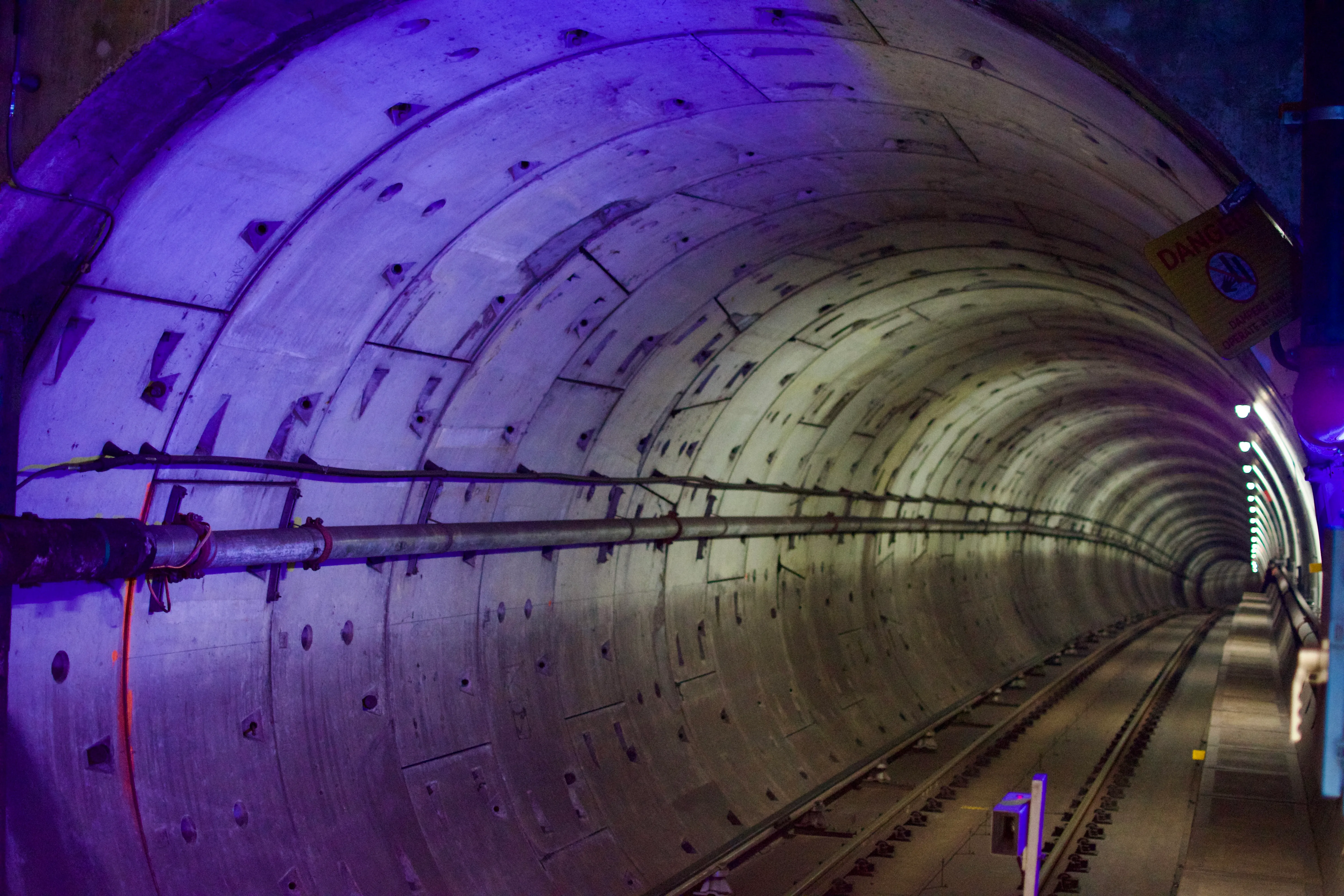 An empty subway tunnel extends into the distance. The closest part of the tunnel is illuminated by a blue LED or a blacklight. The rest of the tunnel has white lights spaced at intervals.