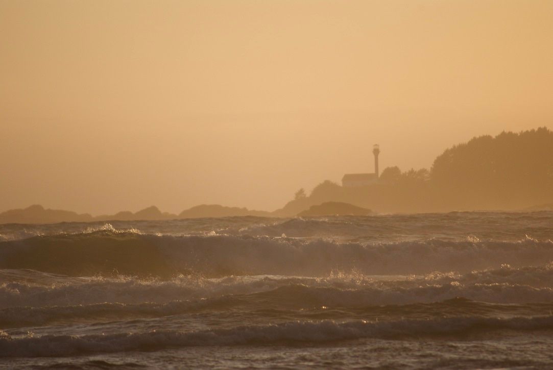 The orange silhouette of a lighthouse building and tower while waves crash onto the beach in the foreground.