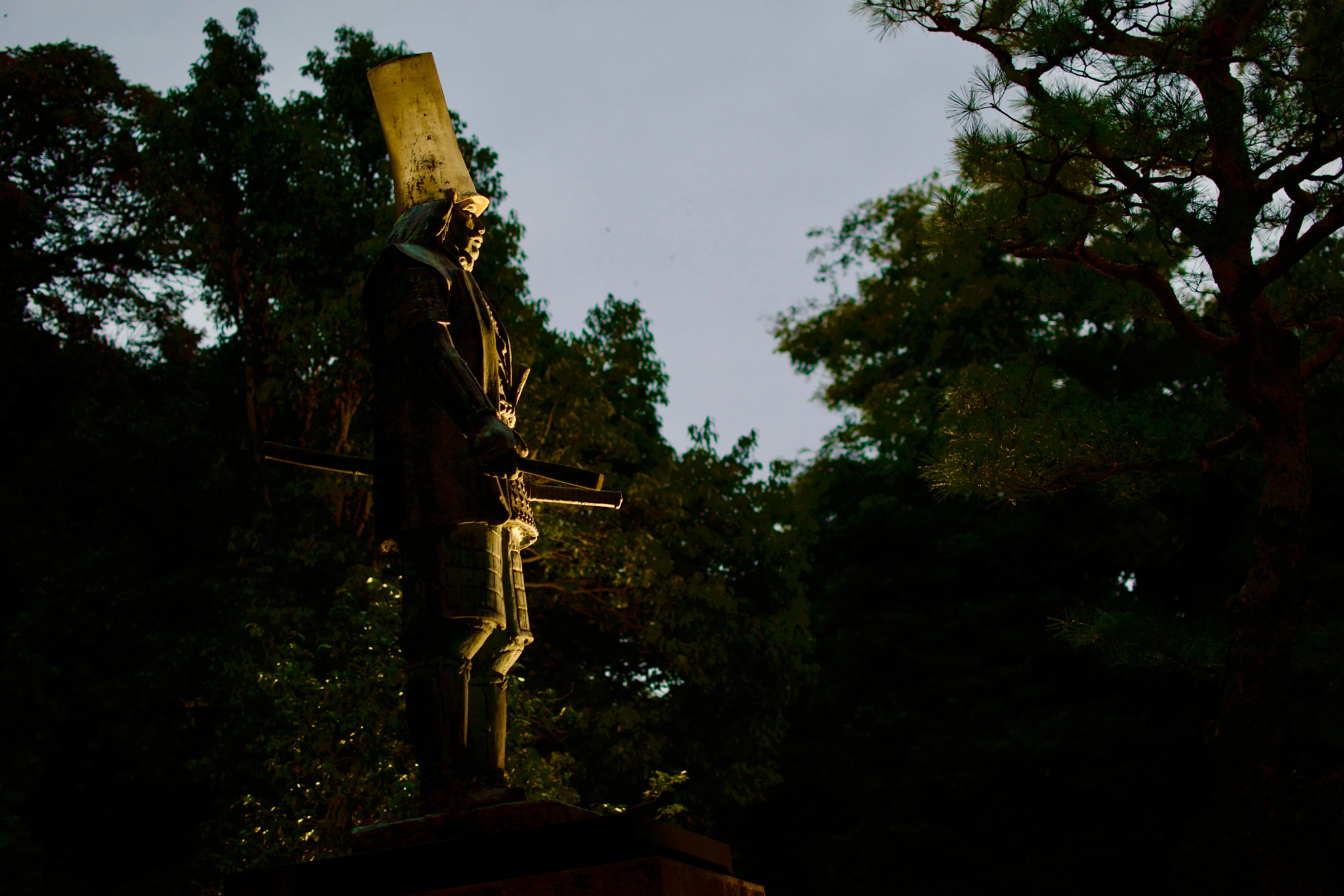 A statue of a samurai with a large gold-plated hat, among dark trees at twilight, dramatically lit from below