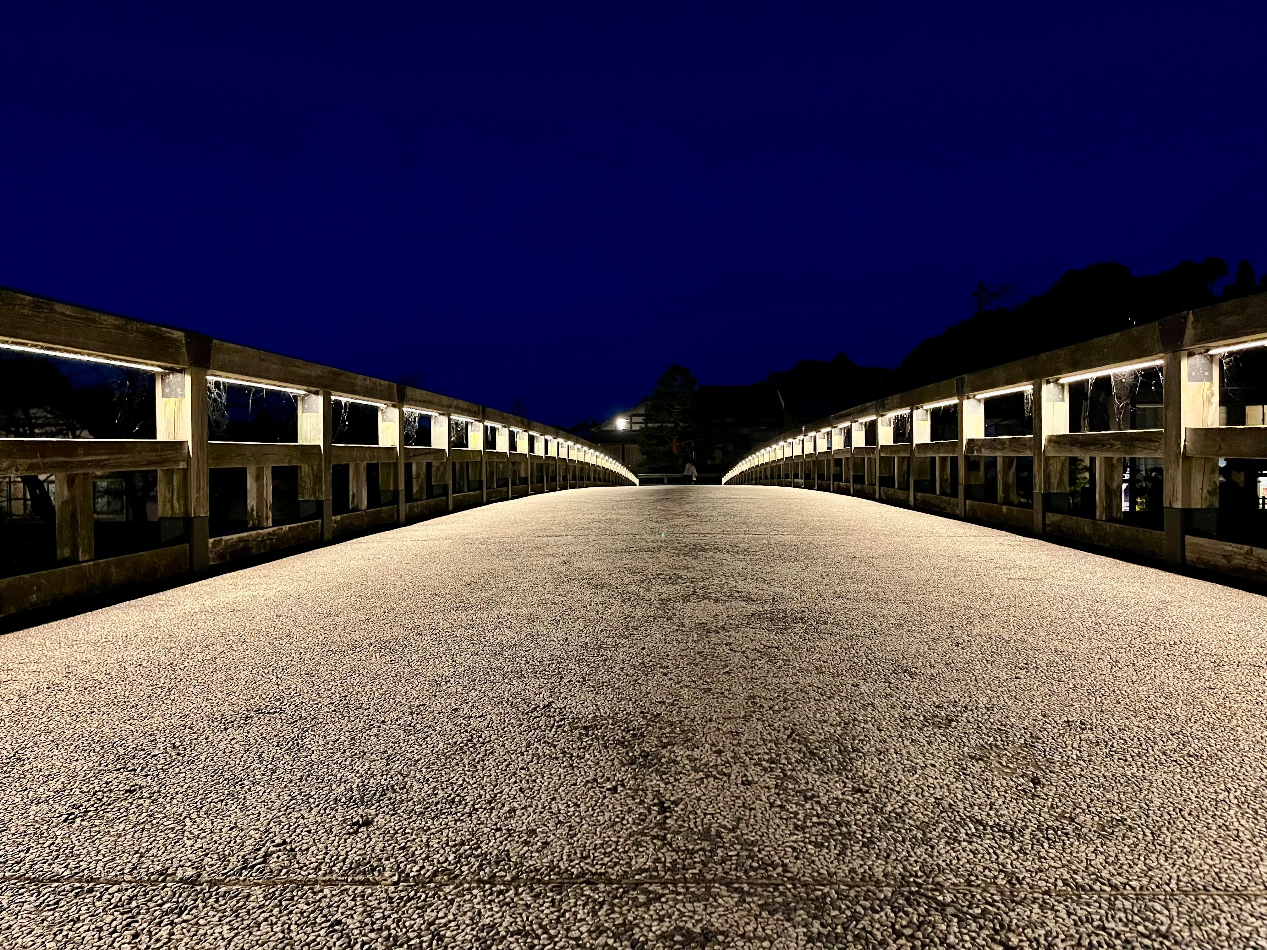 A knee-level view looking across a wooden pedestrian bridge. The bridge is illuminated by lights under the guardrails, and the sky above is a deep navy blue after sunset.