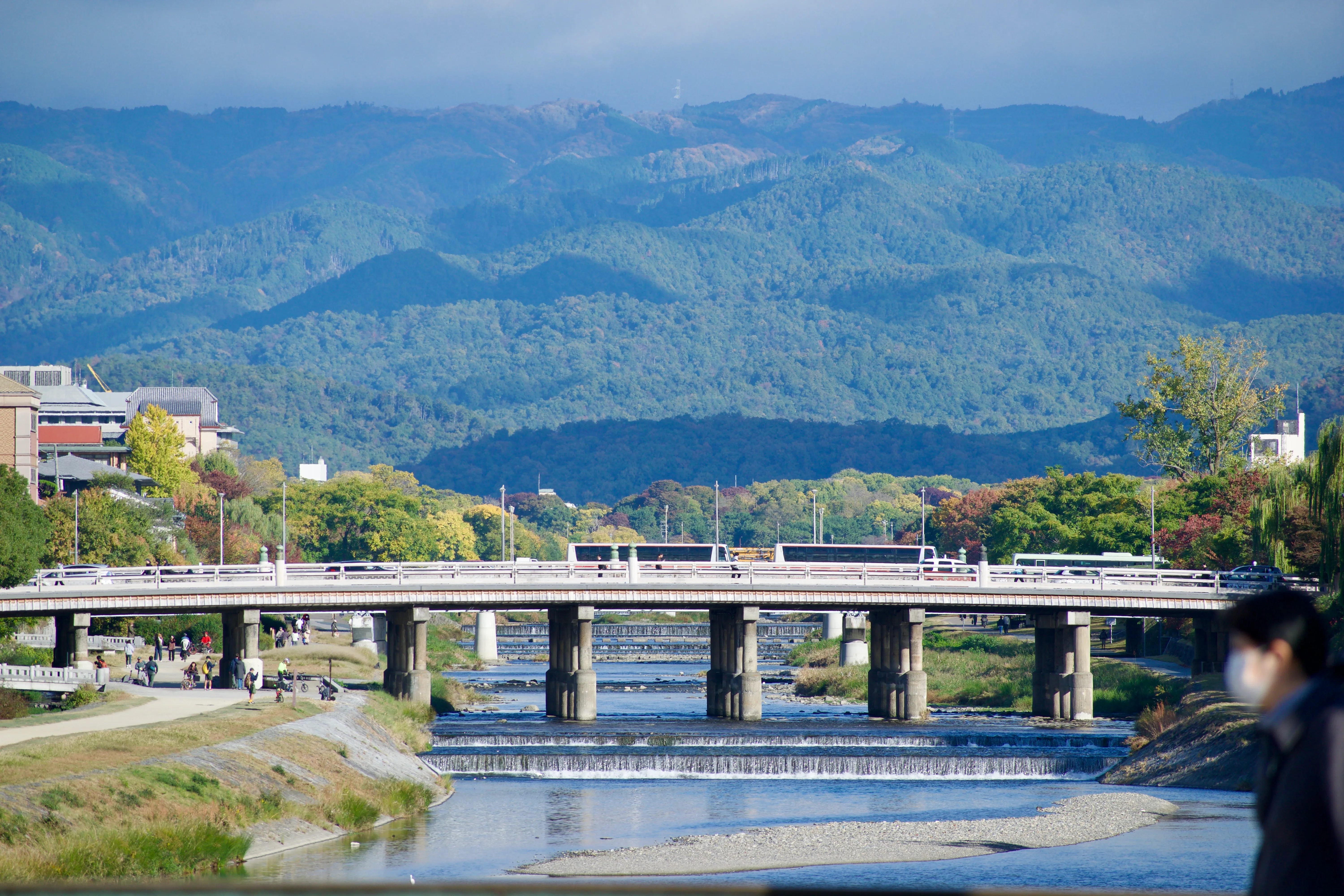 A view of the mountains surrounding Kyōto. In the foreground, a bridge spans the shallow waters of the Kamo river. There are tour buses on the bridge and people walking on the riverbank path to the left.