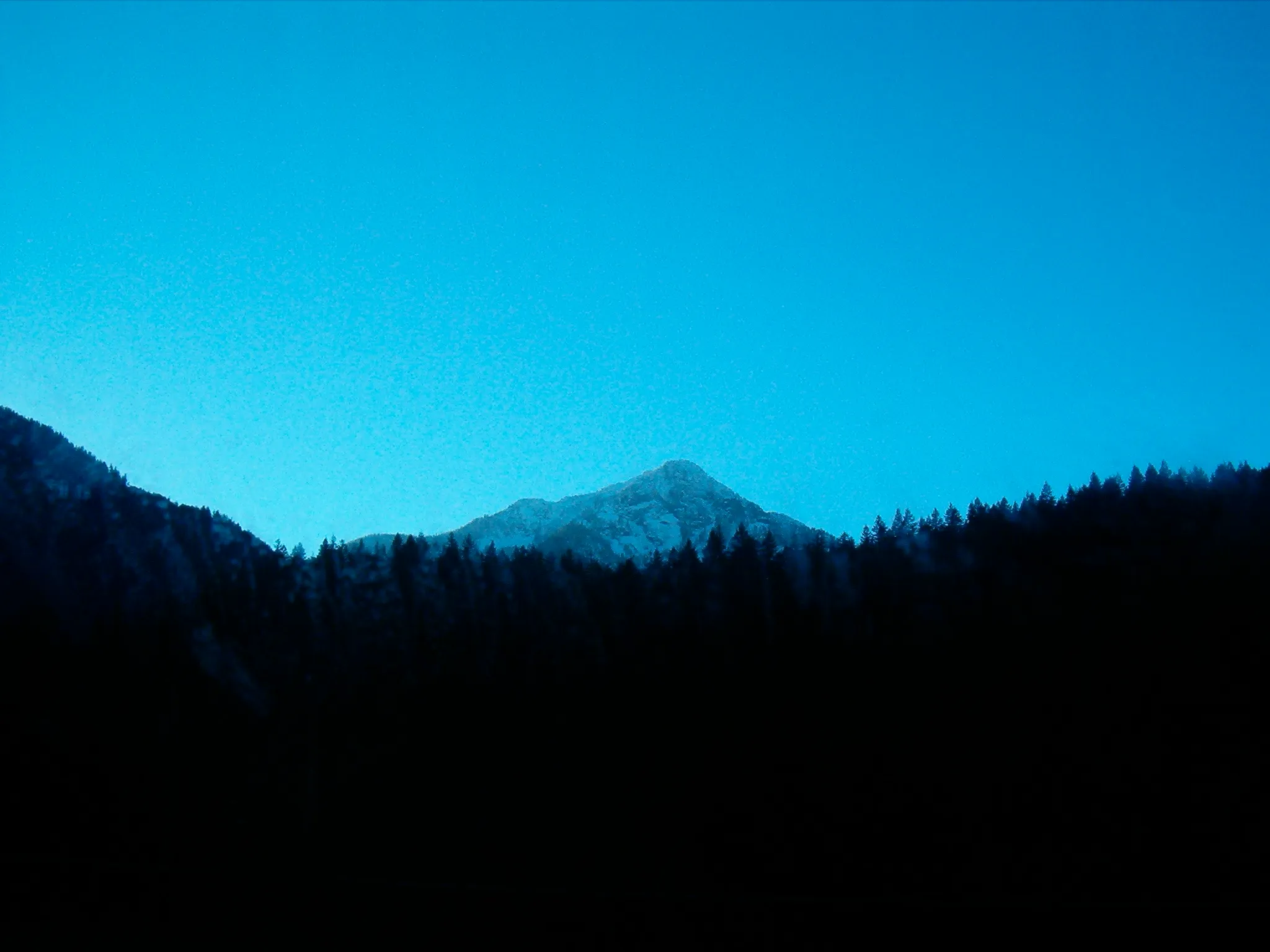 Snow-covered mountain peak rises prominently above a dark, forested landscape. The peak is sharply defined against a dusky sky.
