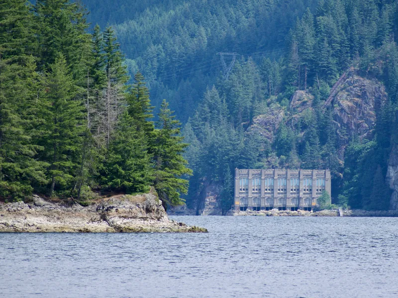 Across the inlet in the distance, a large rectangular concrete building sits on the water at the base of a forested mountain. The building has large windows that have been covered. There is no apparent access to the building, which sits at the bottom of a cliff.