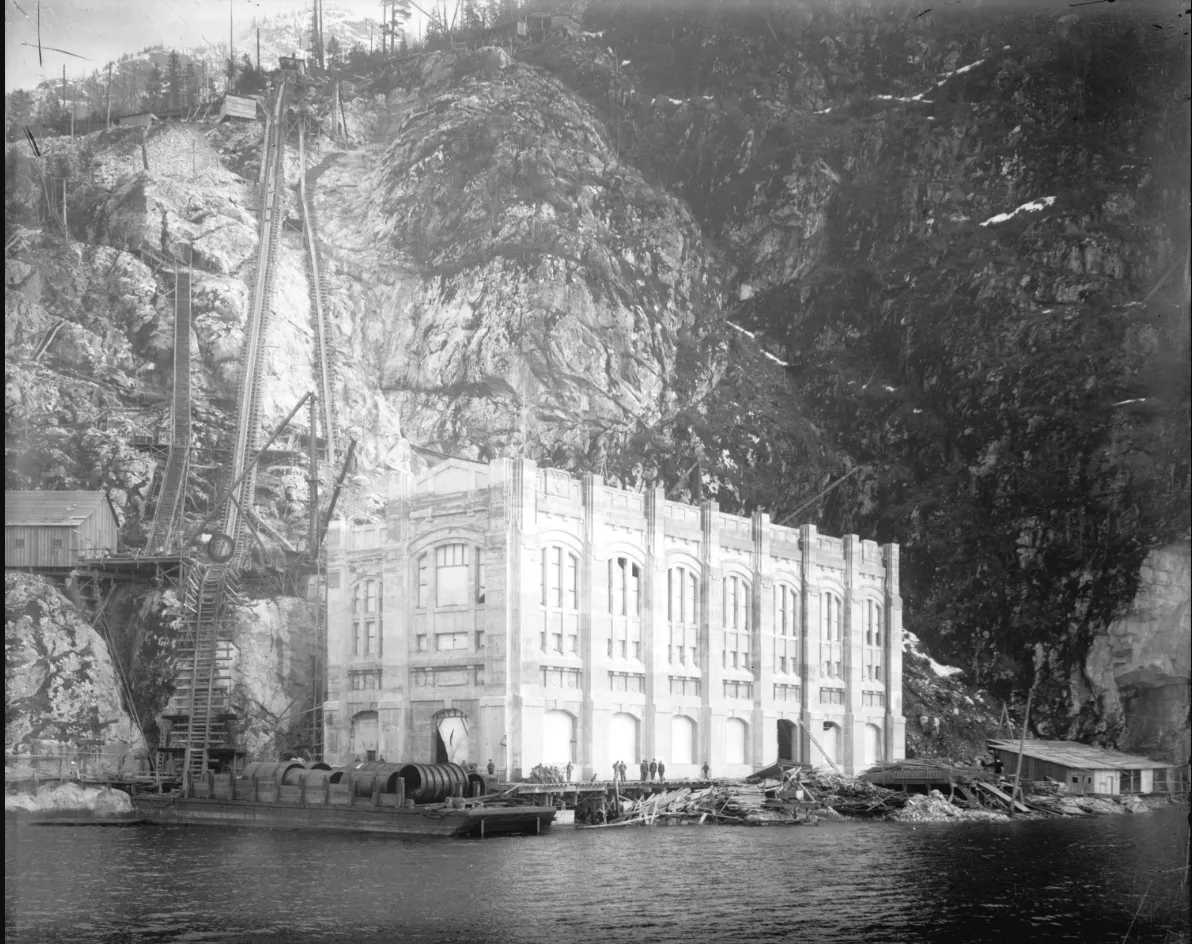 A black-and-white photo of a large but narrow concrete building at the bottom of a steep hill on the water. The exterior is mostly complete, but there are large pipes outside indicating that construction on the inside is ongoing. 