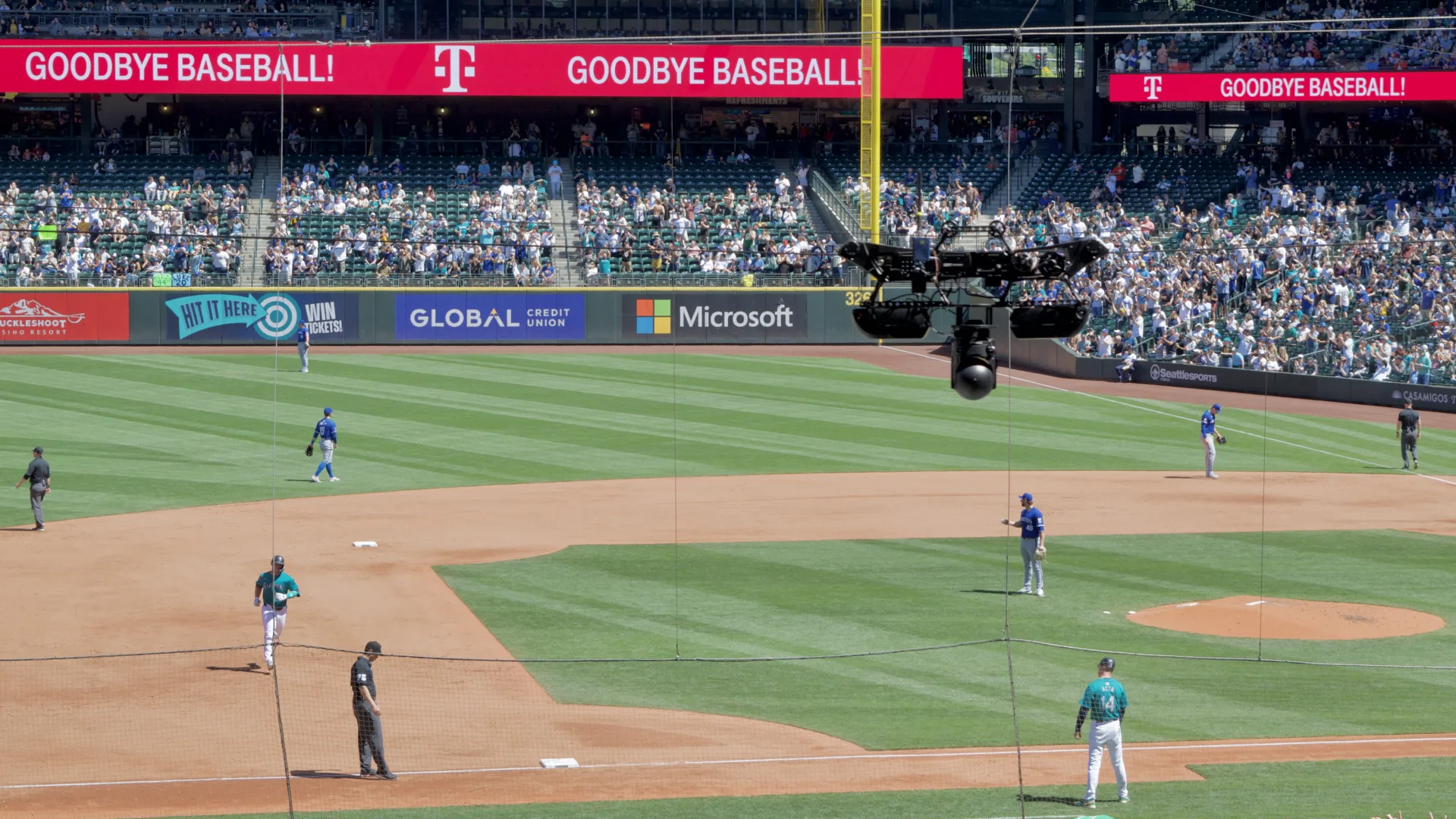 A mobile camera hanging from a pair of cables follows a Seattle Mariners baseball player jogging towards third base.