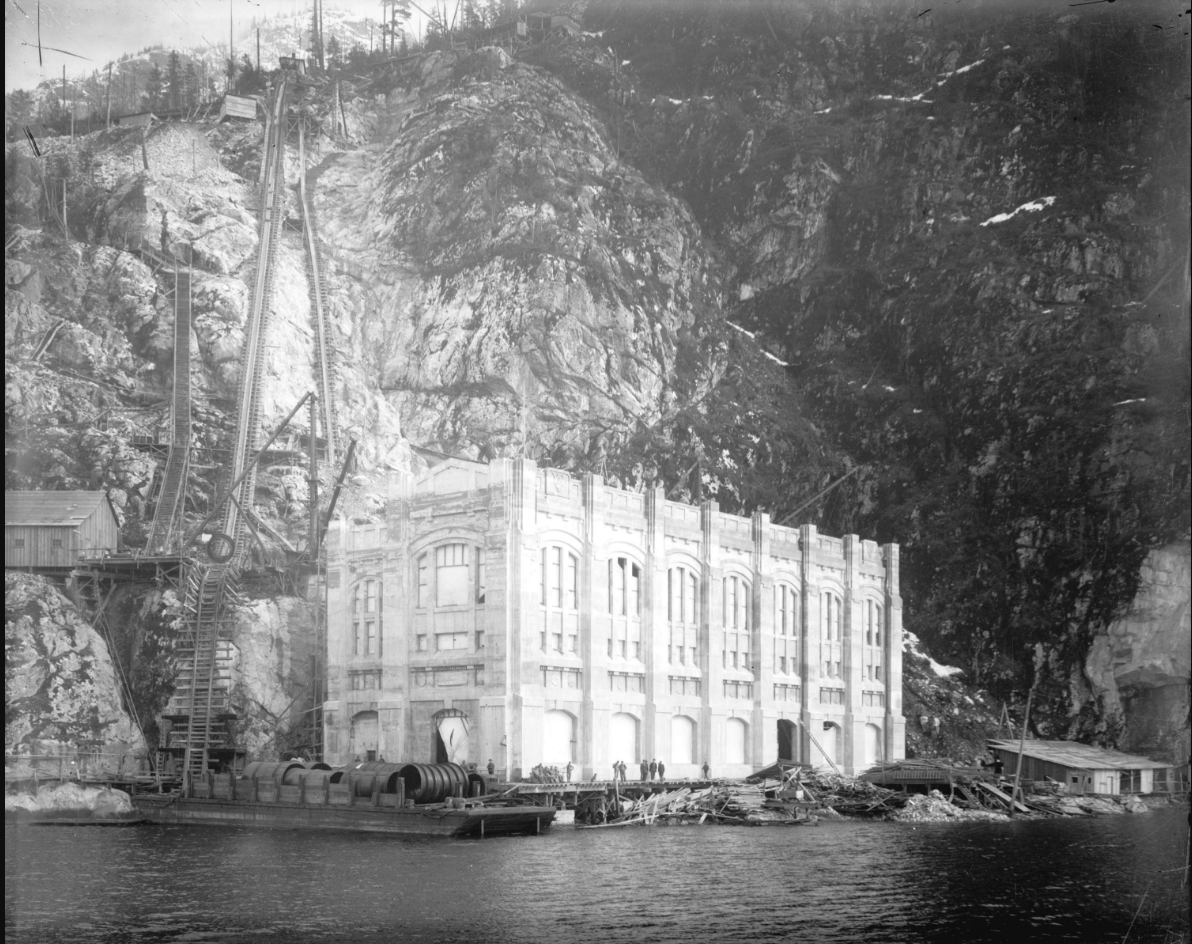 A black-and-white photo of a large but narrow concrete building at the bottom of a steep hill on the water.